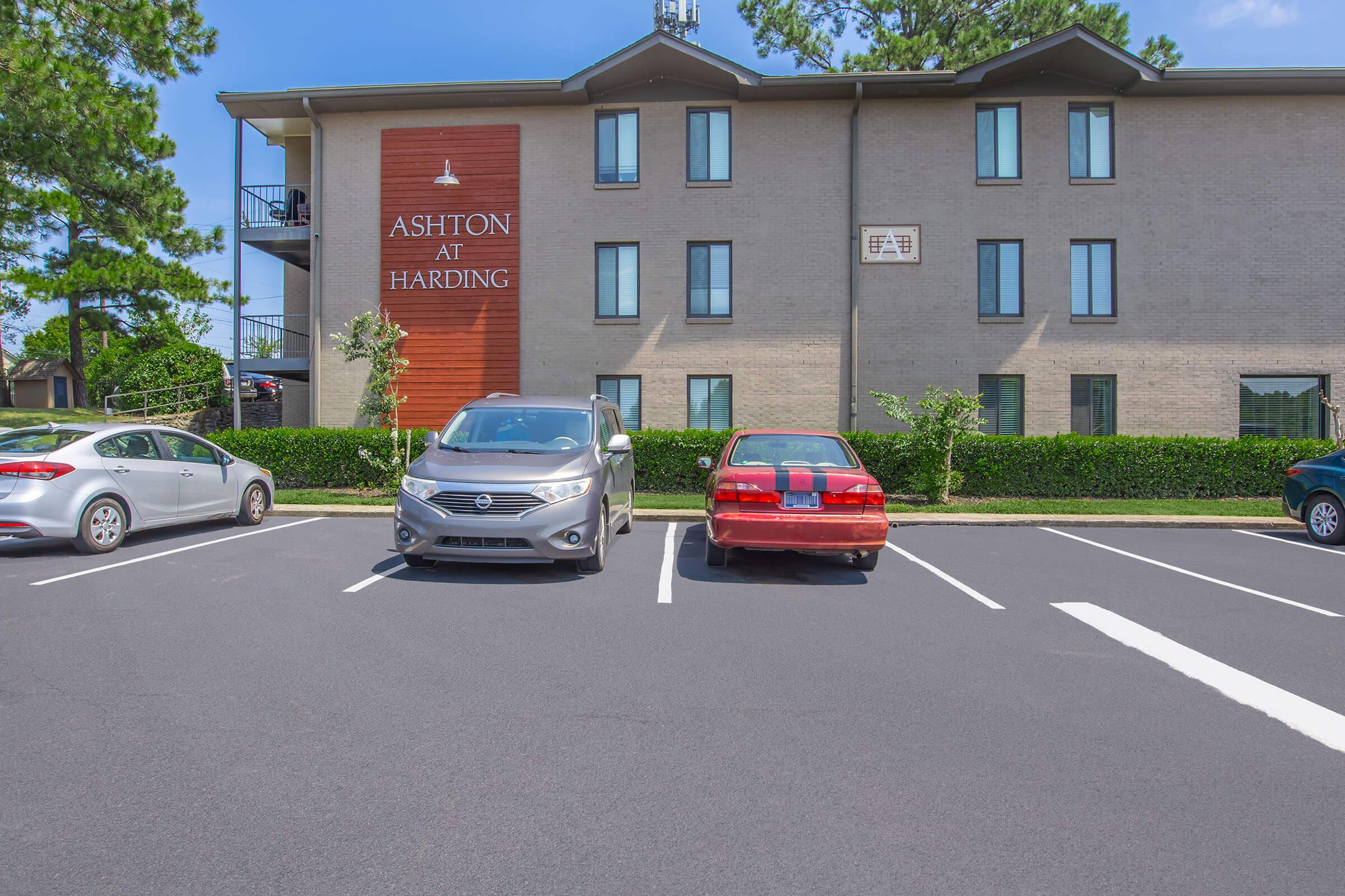 View of the Ashton at Harding apartment building. Two parked cars are visible in the foreground, one silver SUV and one red sedan. The building features a wood-paneled sign prominently displaying the name "Ashton at Harding," with greenery and a well-maintained parking lot surrounding it.