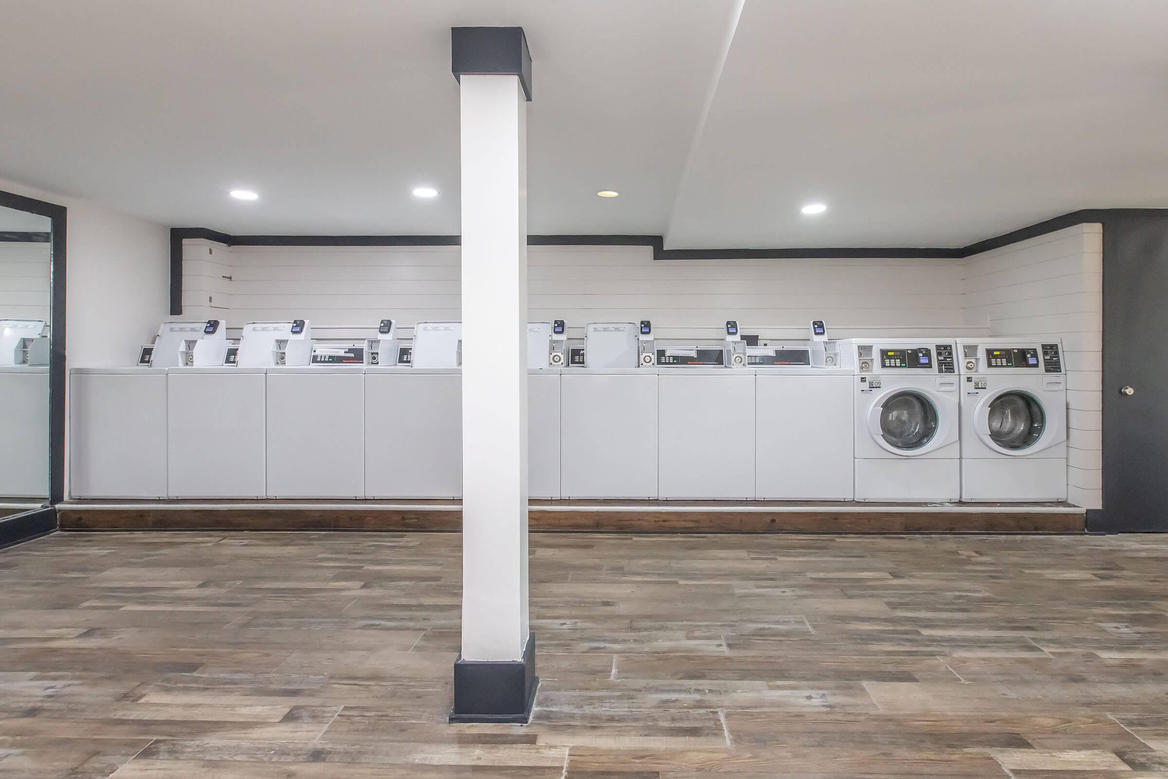 A modern laundry room featuring several stacked white washing machines and dryers aligned against a wall. The space has a clean, minimalist design with wood-like flooring and bright lighting. A central support column divides the area, contributing to the contemporary look.