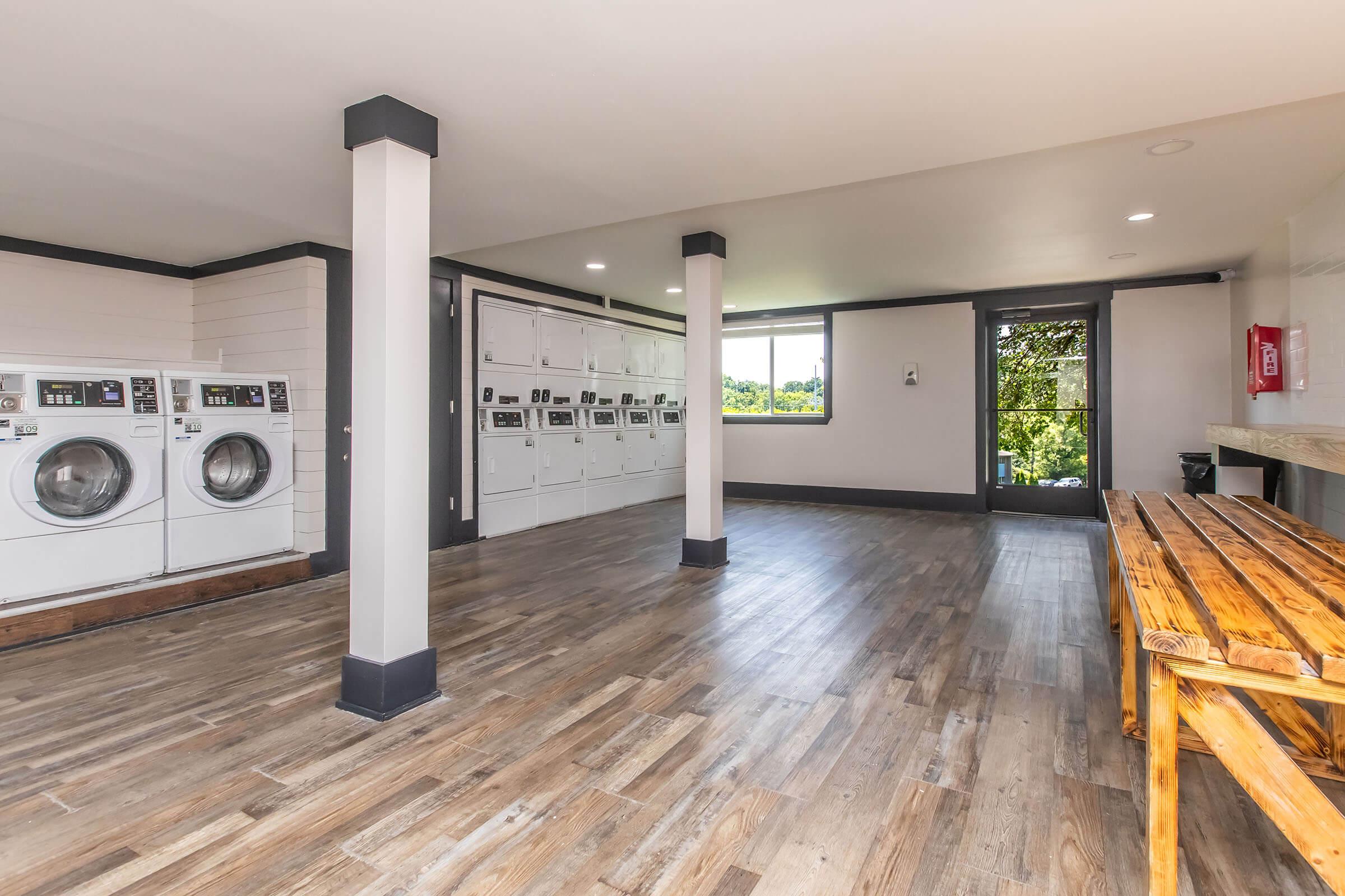 A spacious and modern laundry room featuring several washing machines and dryers, a row of lockers for storage, a large window allowing natural light, and a wooden bench along one wall. The room has a clean, minimalist design with hardwood flooring and light walls.