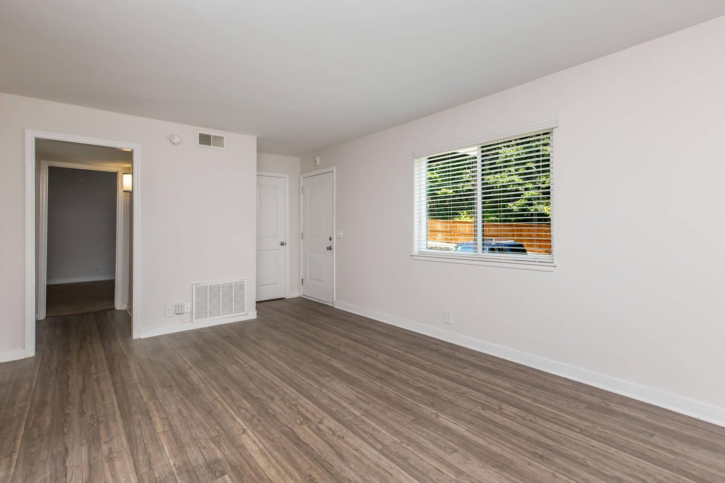 A spacious, empty living room with light-colored walls and hardwood flooring. A window with blinds allows natural light to enter, and there is a door leading to another room. The hallway visible in the background also has a door, contributing to the open and airy feel of the space.