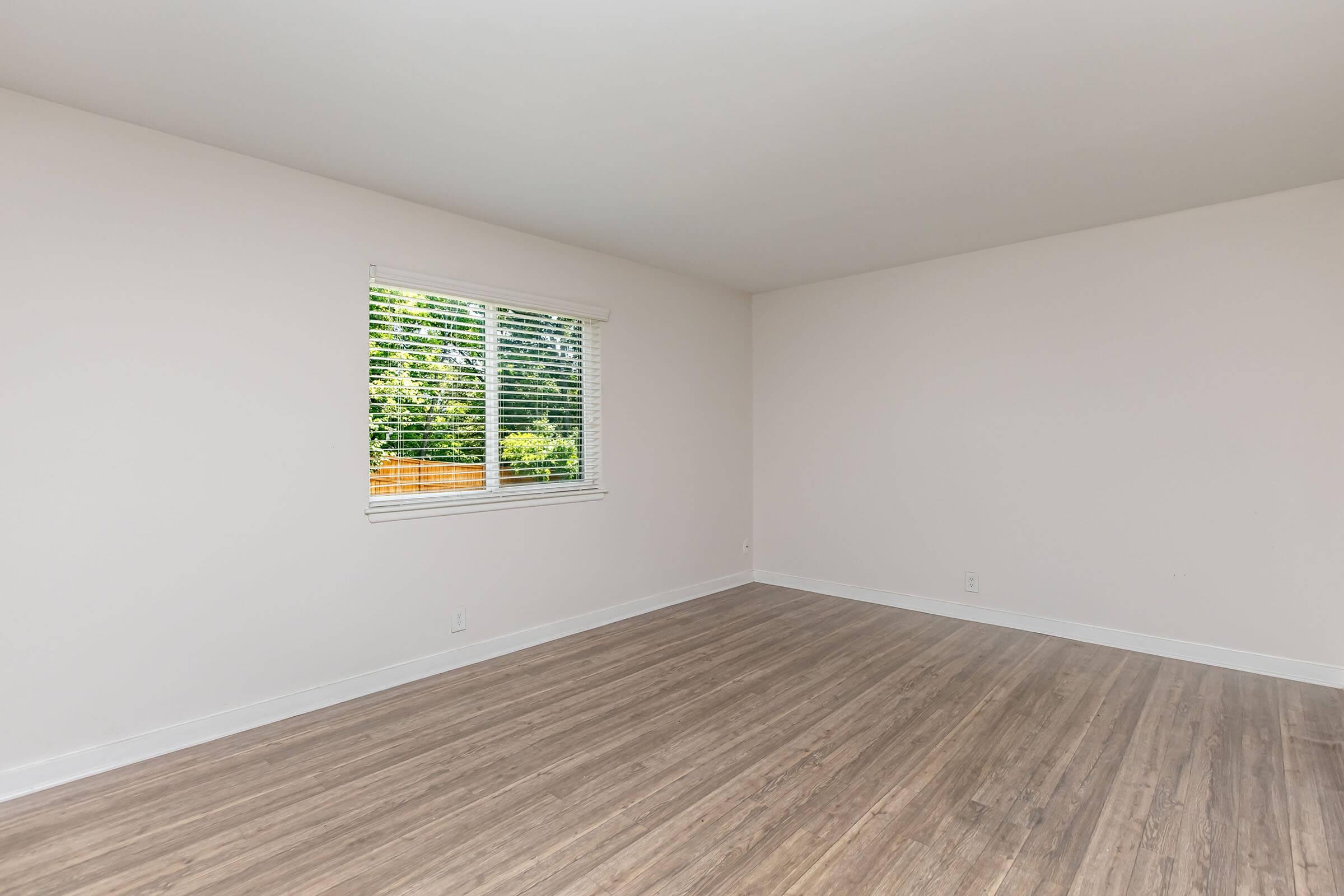 A spacious, empty room with light-colored walls and wooden flooring. There is a window on one wall, allowing natural light to enter, with greenery visible outside. The room is uncluttered and ready for furnishing, creating a bright and welcoming atmosphere.