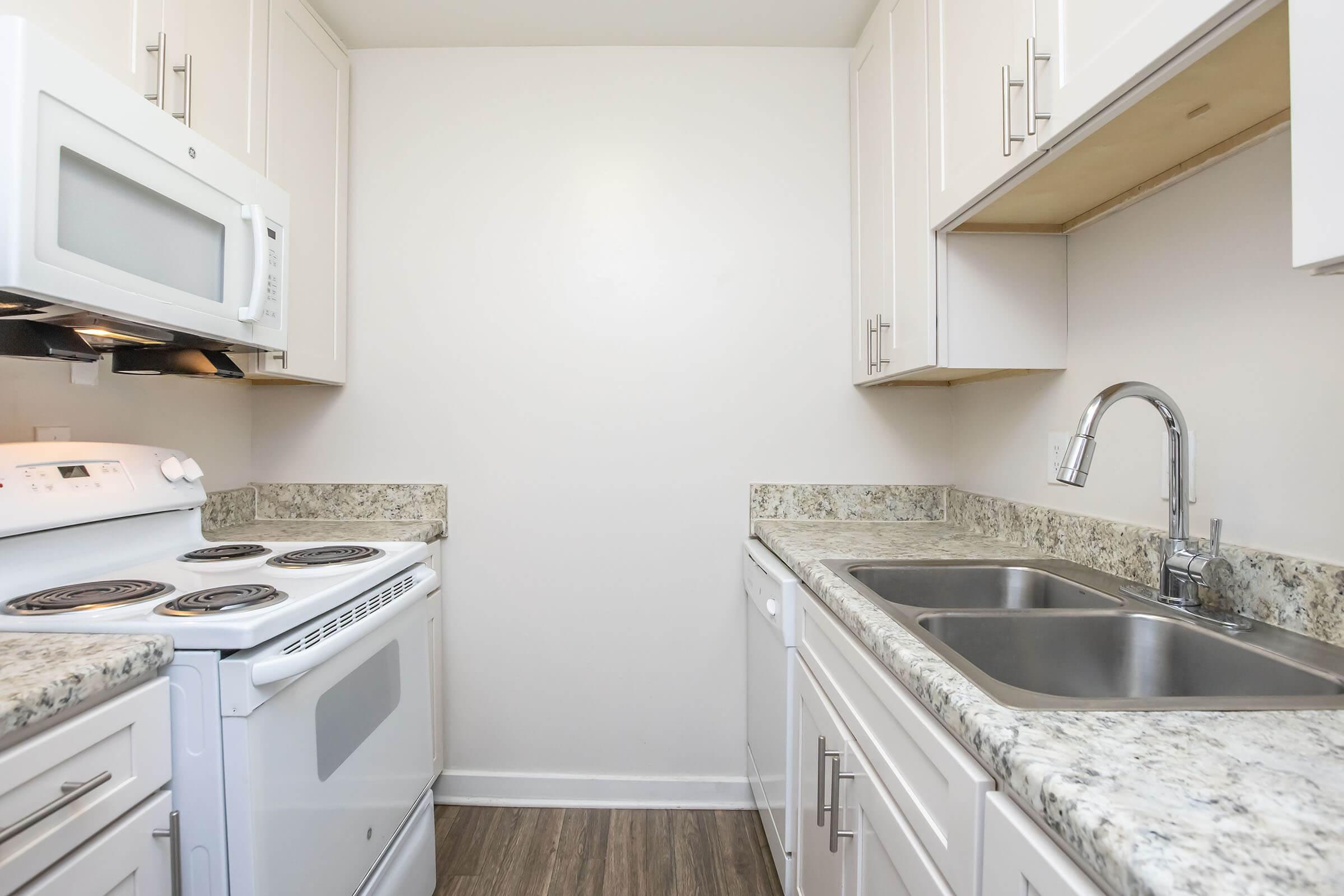 Compact kitchen featuring white cabinets, a granite countertop, a double sink with a modern faucet, an electric stove with an oven, and a microwave mounted above. The flooring is a light wood laminate, and the walls are painted a neutral color, creating a bright and open atmosphere.