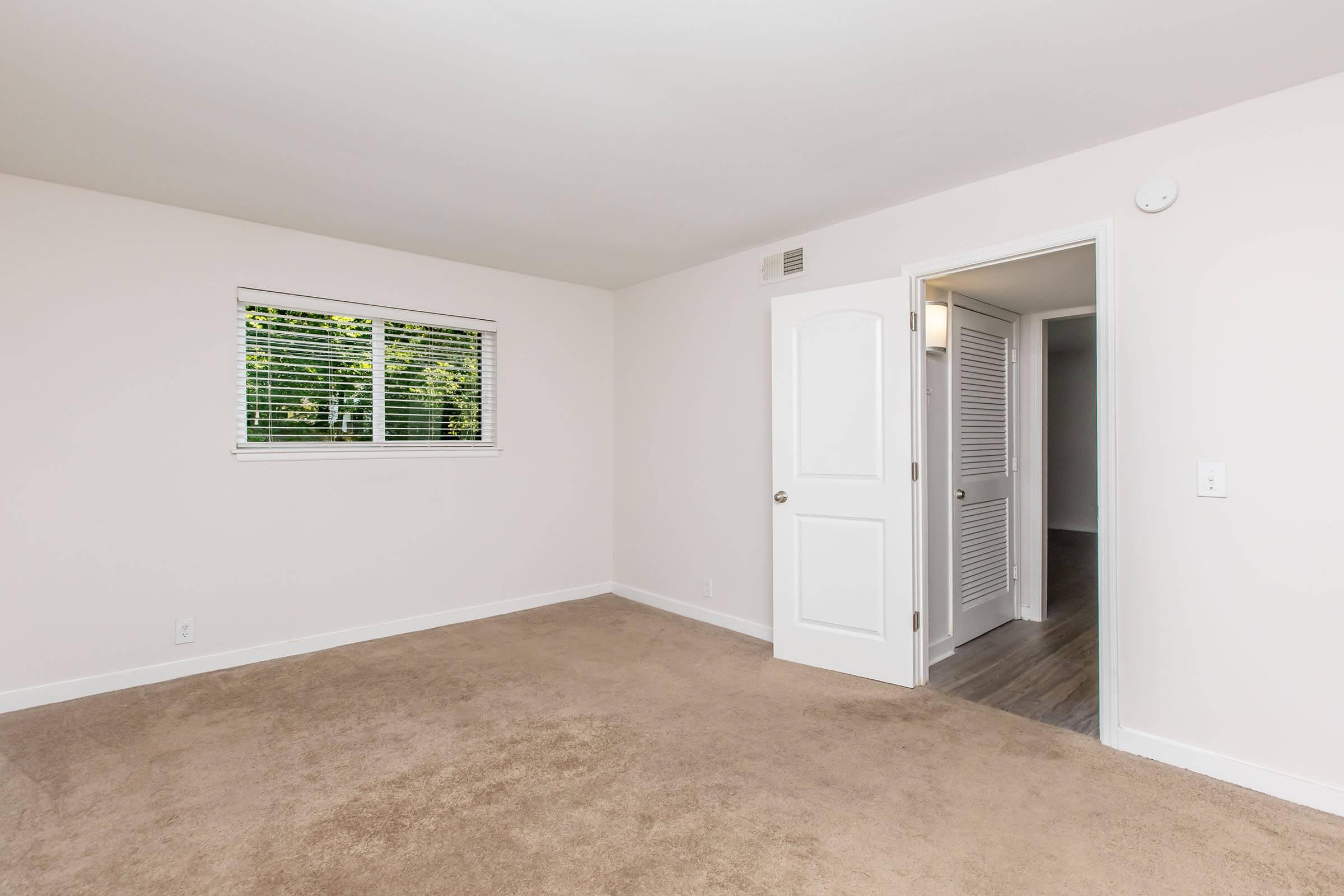 A spacious, light-filled room with beige carpet and white walls. There’s a window with wooden blinds, providing natural light, and a door leading to another room. The overall ambiance is clean and minimalistic, ideal for various uses such as a bedroom or office.