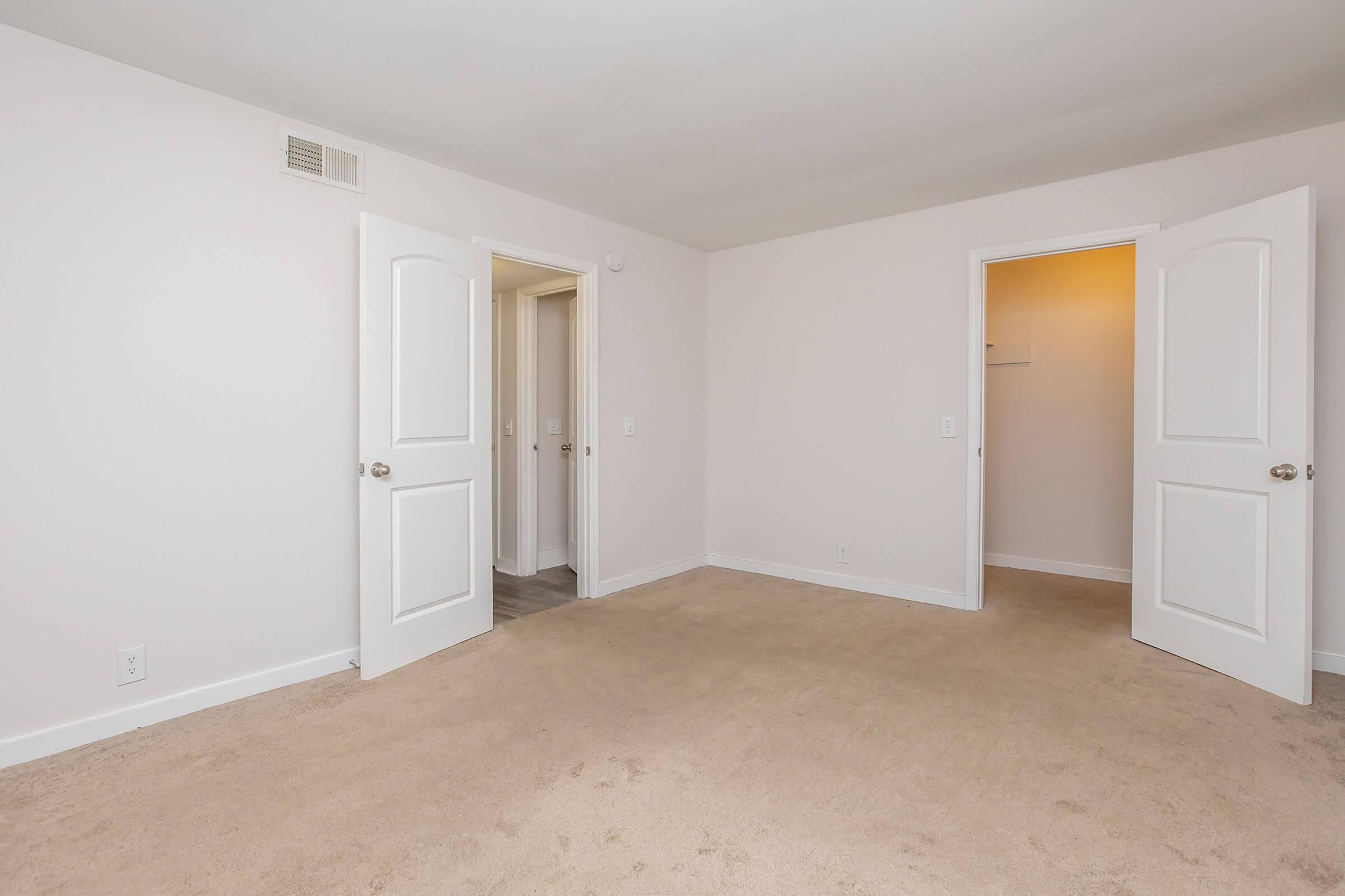 A spacious, empty room with light beige carpet and pale pink walls. Two white doors are open, leading to adjacent spaces, adding to the room's bright and airy feel. The absence of furniture emphasizes the room's potential for decoration and personalization.