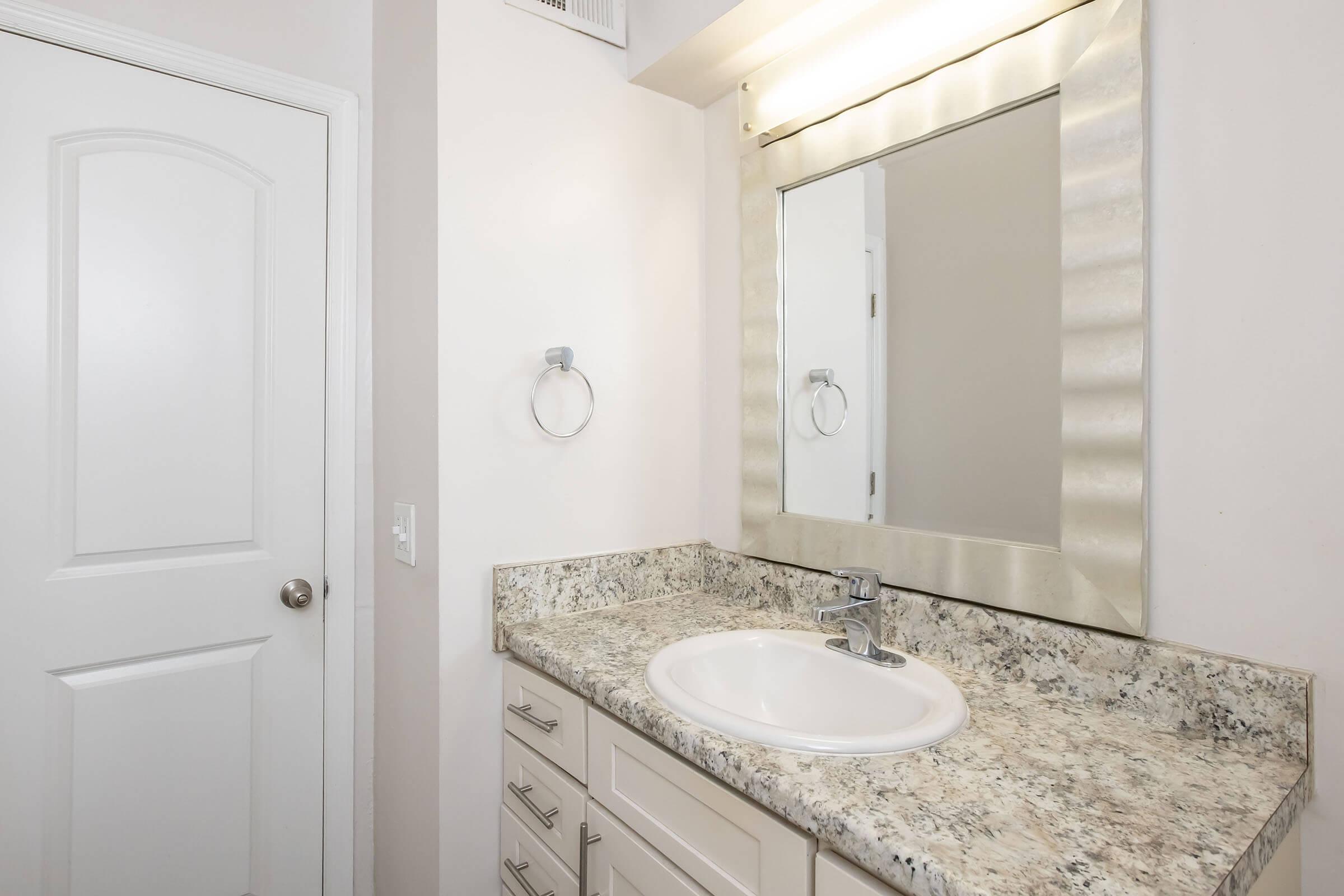 A bright and modern bathroom with light-colored walls. It features a white sink with a polished chrome faucet, a granite countertop, and a large, decorative mirror with a wavy border. There is a towel ring beside the sink and a closed door on the left, adding to the tidy appearance of the space.