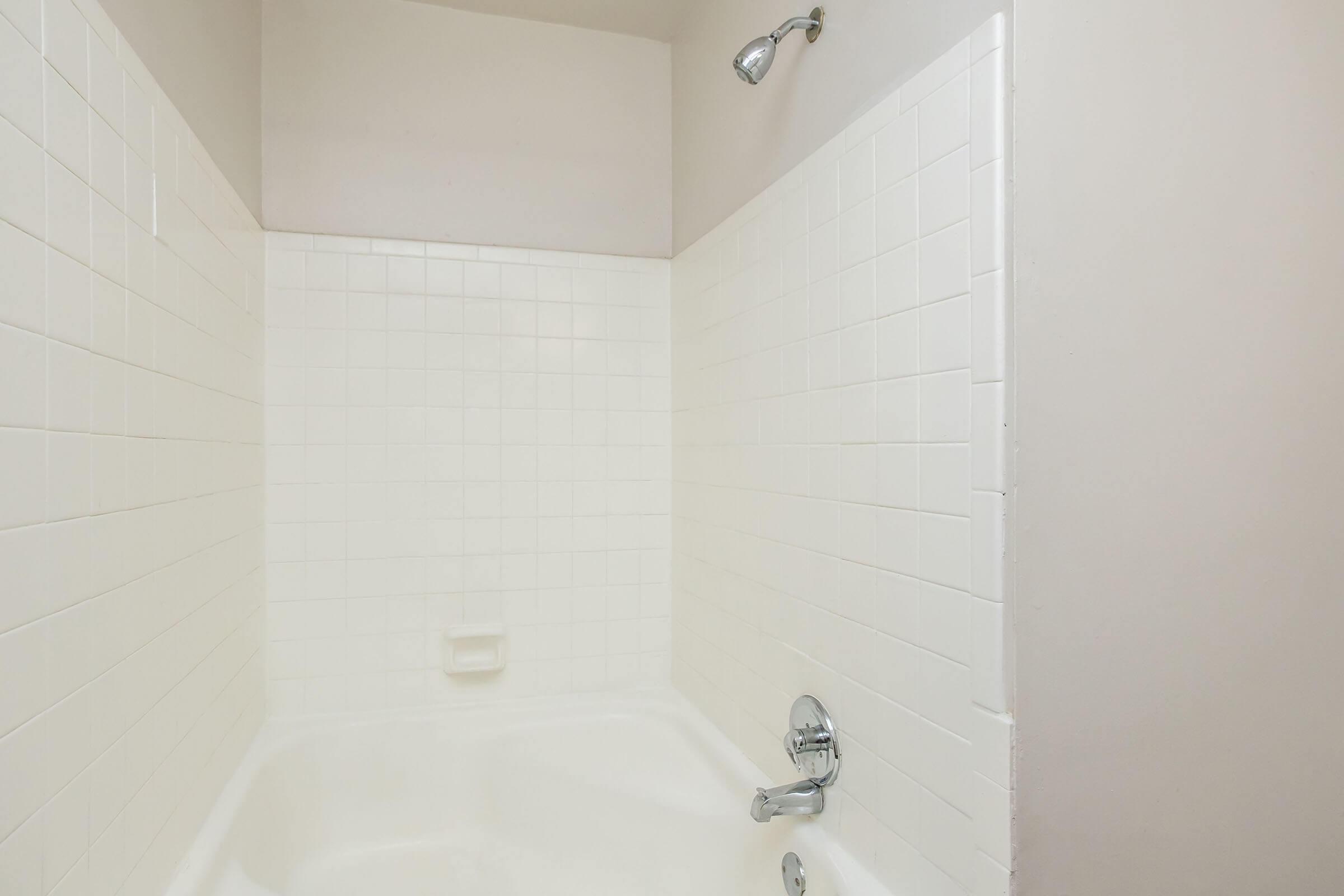 A clean and simple bathroom with a white tiled shower area and a bathtub. The walls are smooth and light-colored, with a showerhead mounted on the wall. The tub has a faucet and shower controls visible, creating a functional and minimalist design.