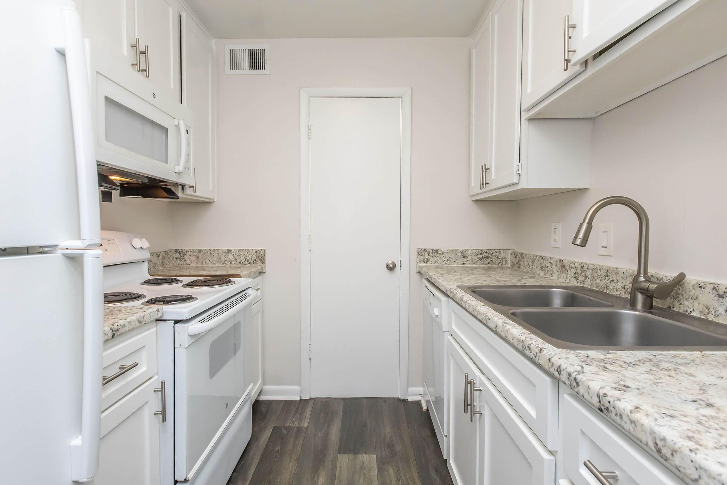 A tidy kitchen featuring white cabinetry, a countertop with a granite finish, a stainless steel sink, and an electric stove. The space is well-lit, with a door at the end leading out of the kitchen area, and modern appliances including a microwave and refrigerator.