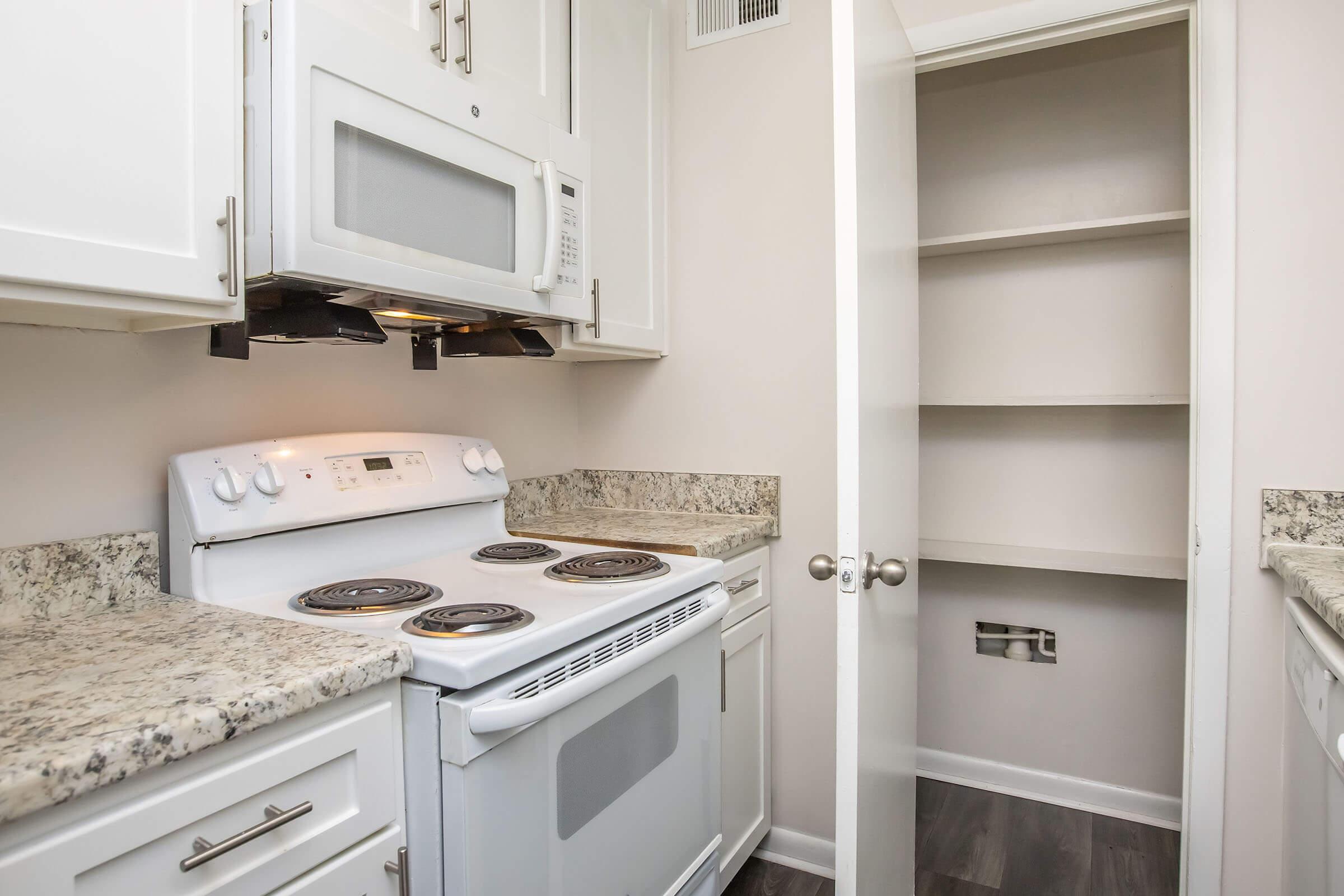 A modern kitchen featuring a white stove and oven, a microwave above, and granite countertops. Cabinets in white finish line the walls, and a nearby door leads to a small pantry with empty shelves. The floor has a dark wood-style finish.