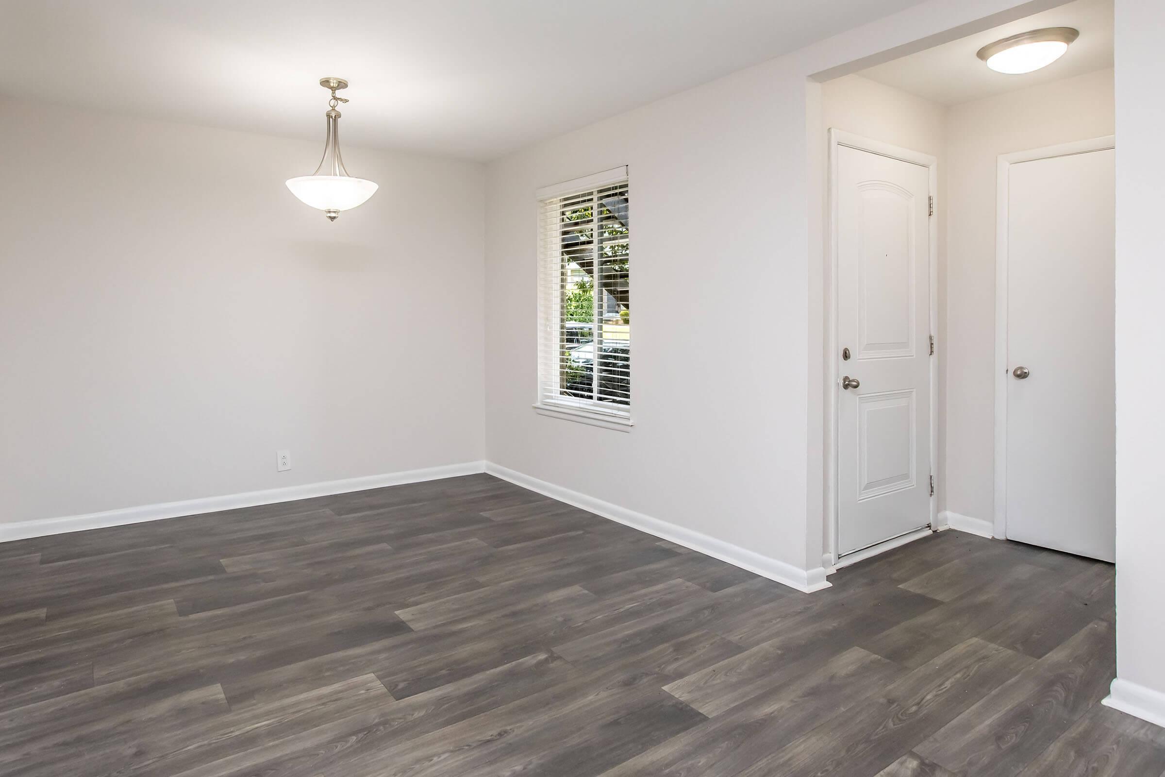 A bright, empty room with light gray walls and new, dark wood laminate flooring. A ceiling light fixture hangs in the center, and there is a window with closed blinds allowing natural light to enter. A white door is visible on the right, leading to another space. The overall atmosphere is clean and modern.