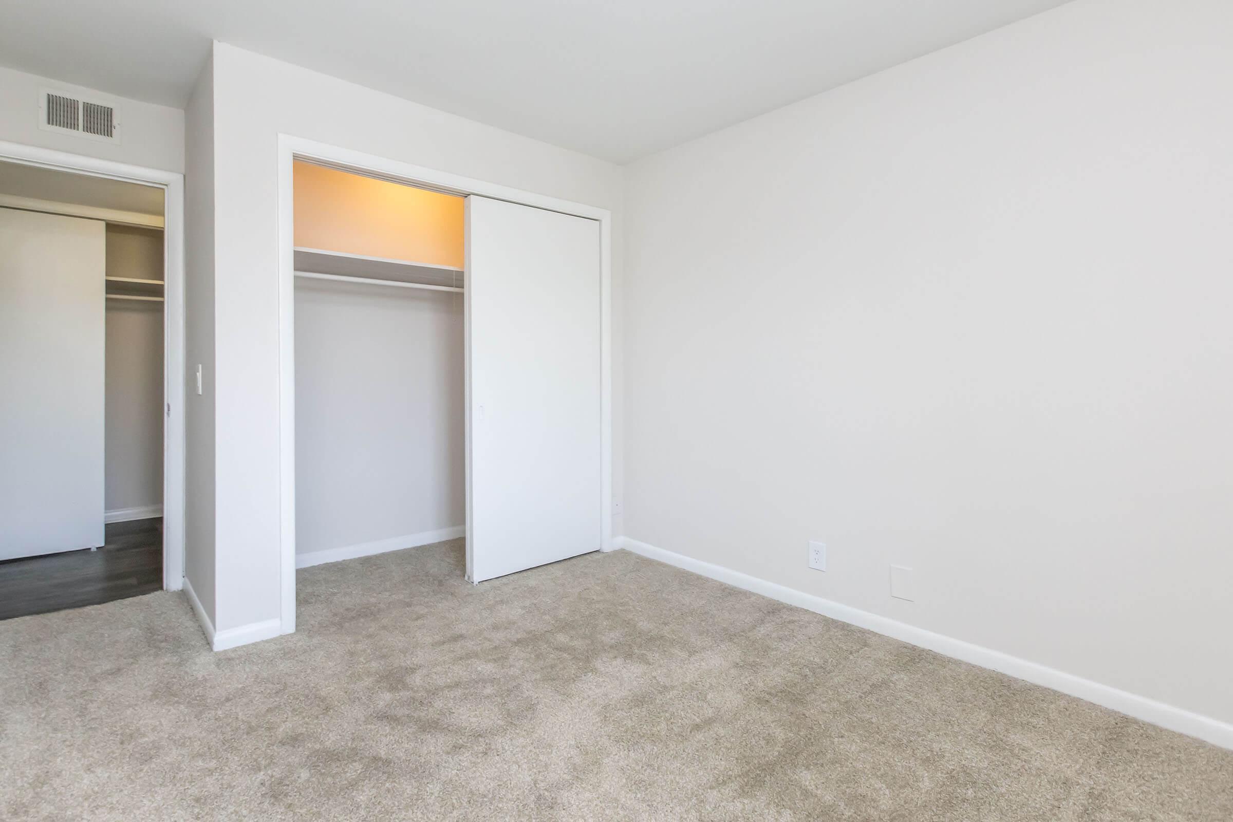 A spacious, empty bedroom featuring light-colored walls and a soft carpet. An open closet with sliding doors is visible on the left side, and the room has ample natural light, creating a bright and airy atmosphere. The floor is clean and uncluttered, emphasizing the room's potential for personalization.