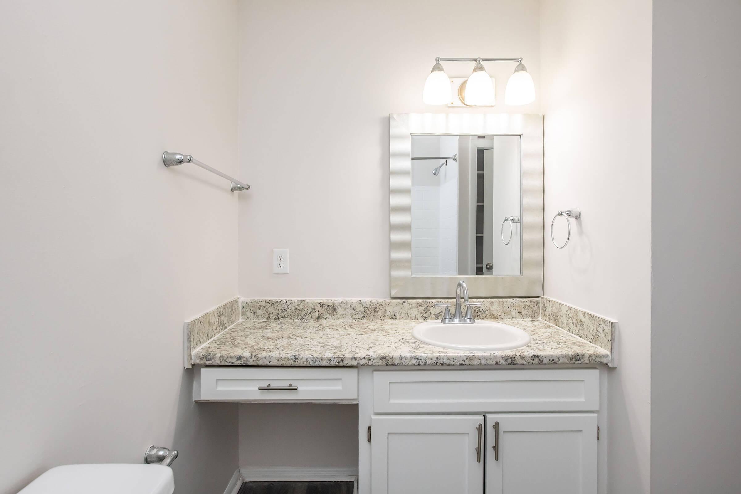 A well-lit bathroom featuring a vanity with a granite countertop, a white sink, a large mirror with three light fixtures above, and a towel rack. The walls are painted in a light color, and there's a toilet visible on the left side. The floor has a dark finish, and a shower can be seen through a door in the background.