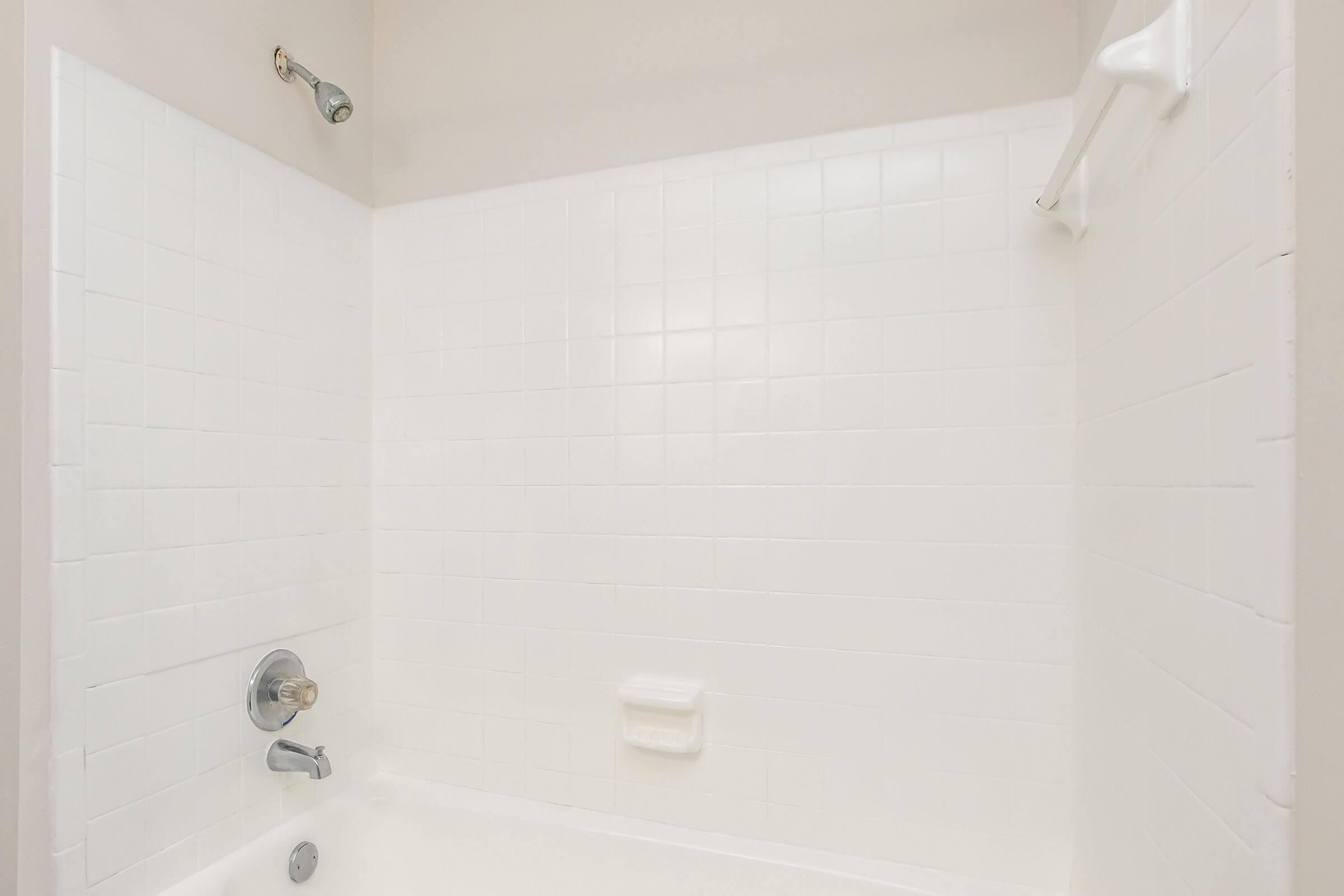 A clean, white tiled shower with a bathtub. The walls are tiled in plain white, featuring a showerhead, a faucet, and a soap dish. The overall ambiance is bright and minimalistic, with no visible decorations or personal items.