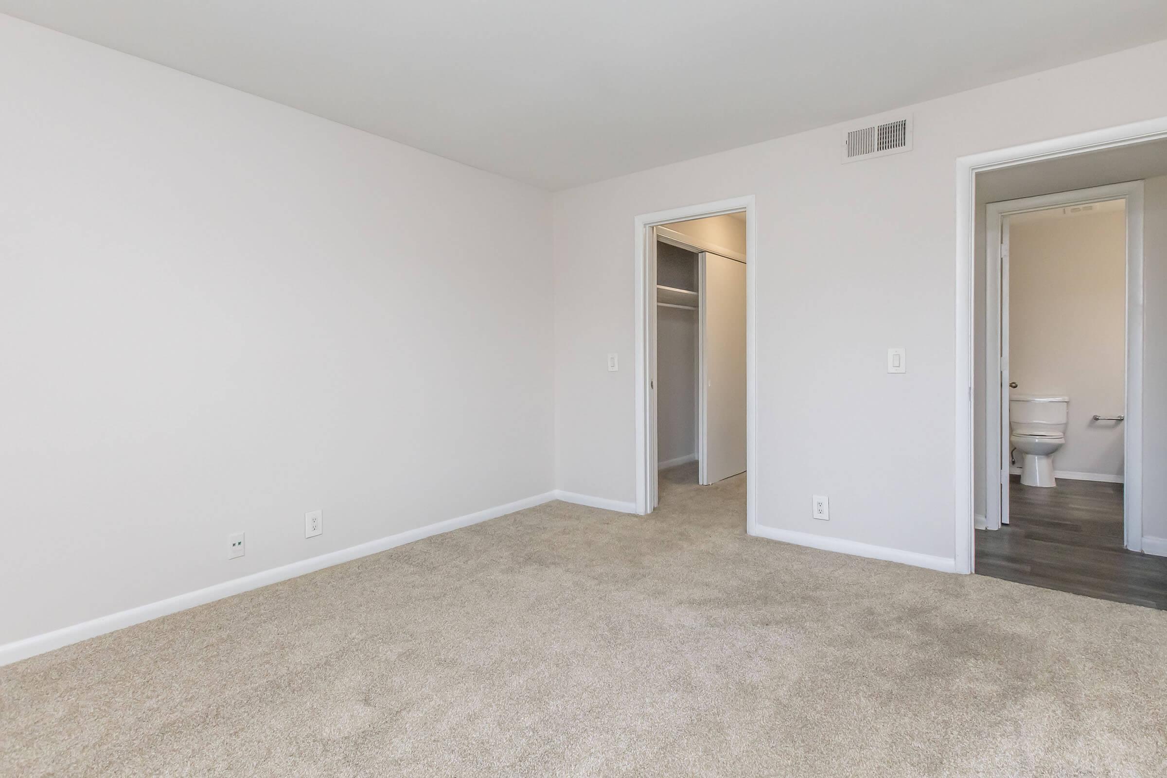 A spacious, empty room with light beige carpet and white walls. There are two doorways leading to adjacent areas; one doorway opens to a closet, and the other leads to a bathroom. The room is well-lit with natural light, creating a clean and neutral space suitable for various uses.