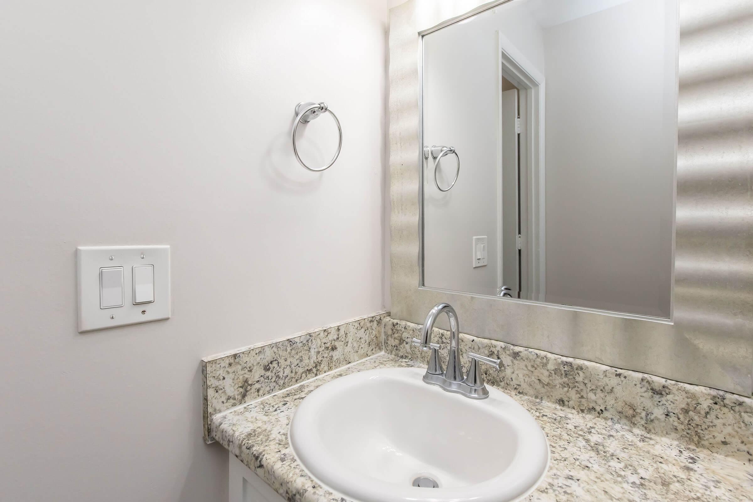 A bathroom sink with a granite countertop, a modern faucet, and a round mirror above it. The wall is painted a light color, and there are light switches on the left side of the image. A towel ring is mounted next to the mirror, and the reflection shows part of a door leading to another room.