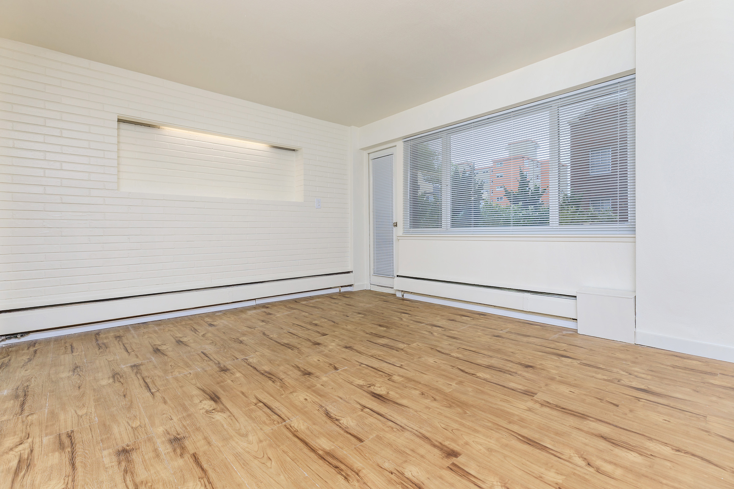 A spacious, empty room featuring light-colored walls and a large window that allows natural light to enter. The floor is made of light wood, and there is a small alcove in one wall. A door leads to another area, and the scene conveys a clean and modern living space ready for decoration.