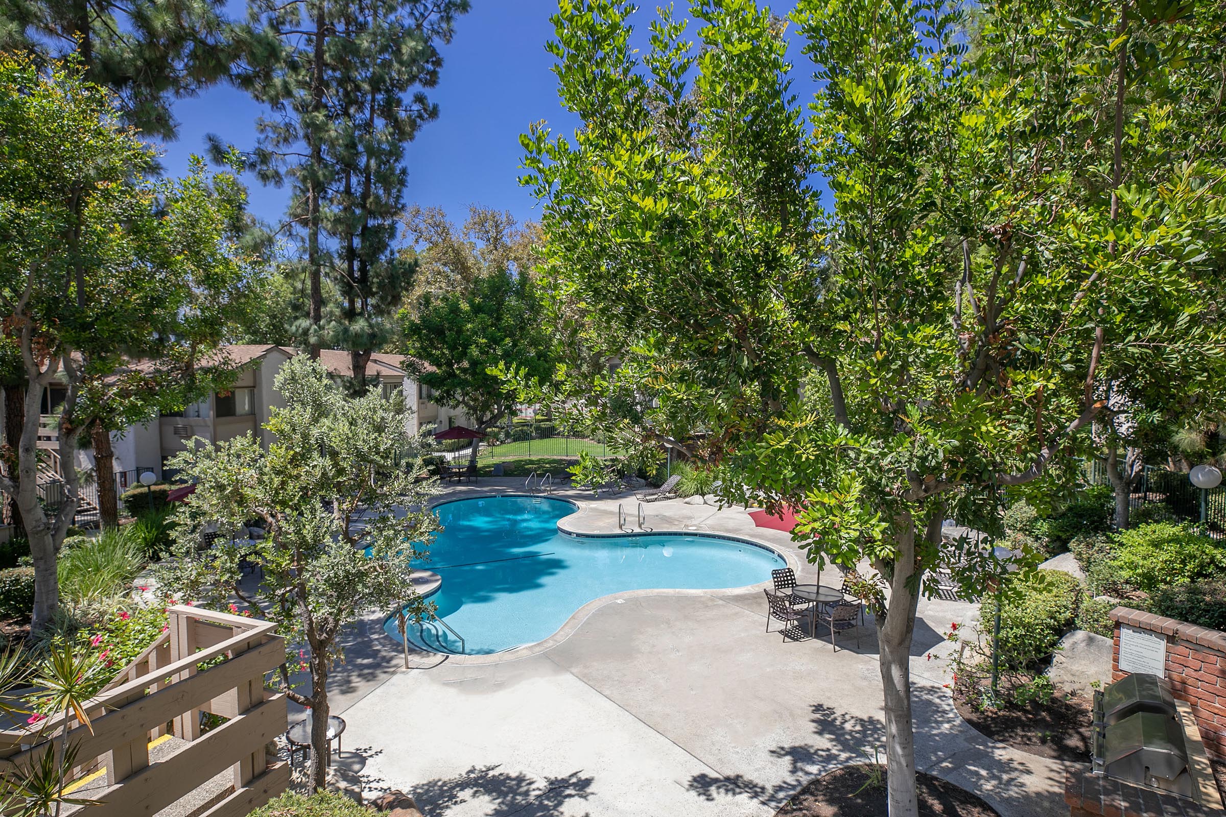 A peaceful outdoor scene featuring a clear blue swimming pool surrounded by lush greenery and trees. Sunlight filters through the leaves, creating a bright atmosphere. There are lounge chairs near the pool and well-maintained landscaping, suggesting a relaxing communal area in a residential complex.