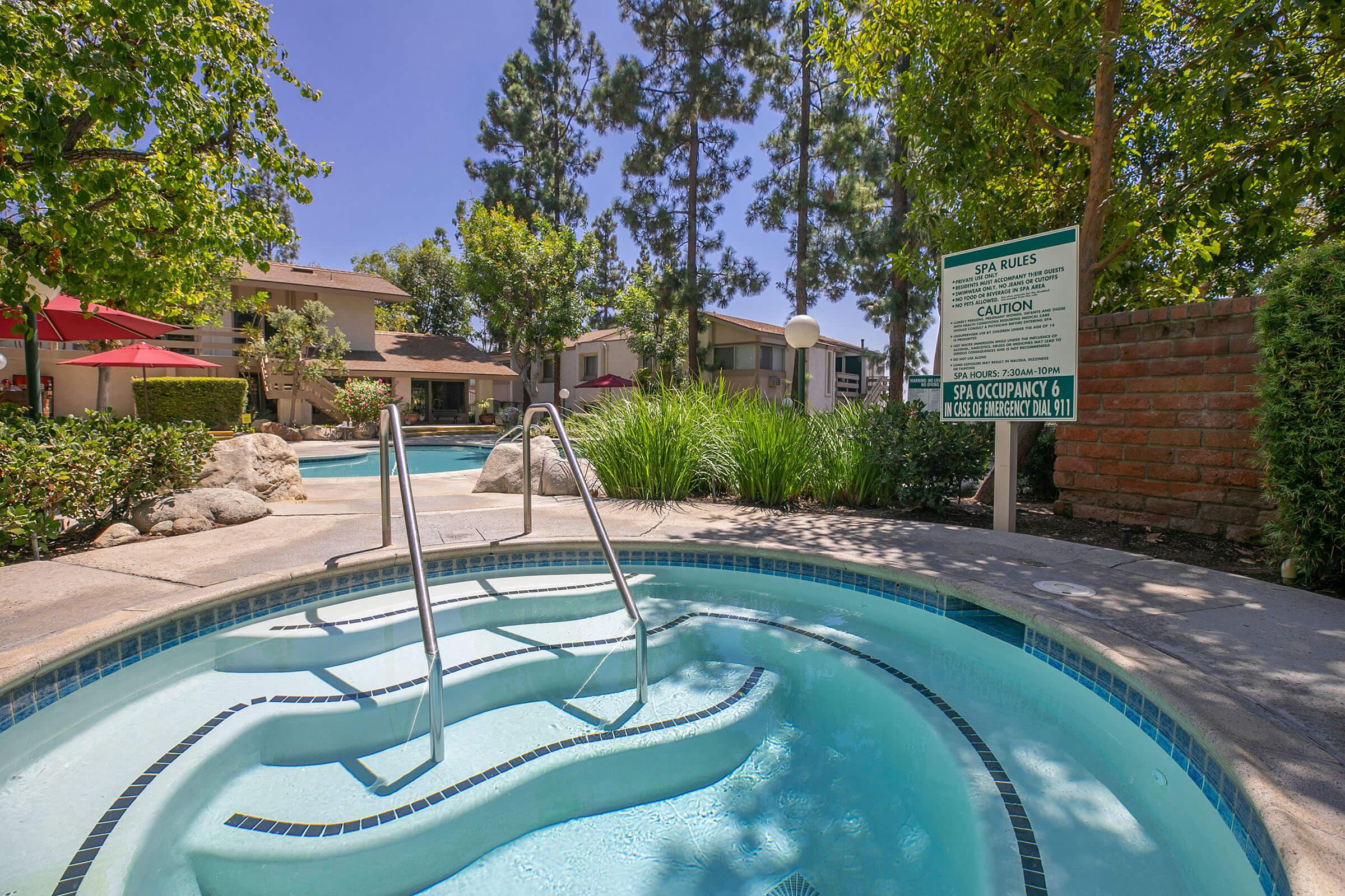 A hot tub with white water and steps leads into it, surrounded by lush greenery and tall trees. In the background, there are residential buildings, and a sign outlining spa rules is positioned nearby. The area is sunny, featuring red umbrellas over seating areas.