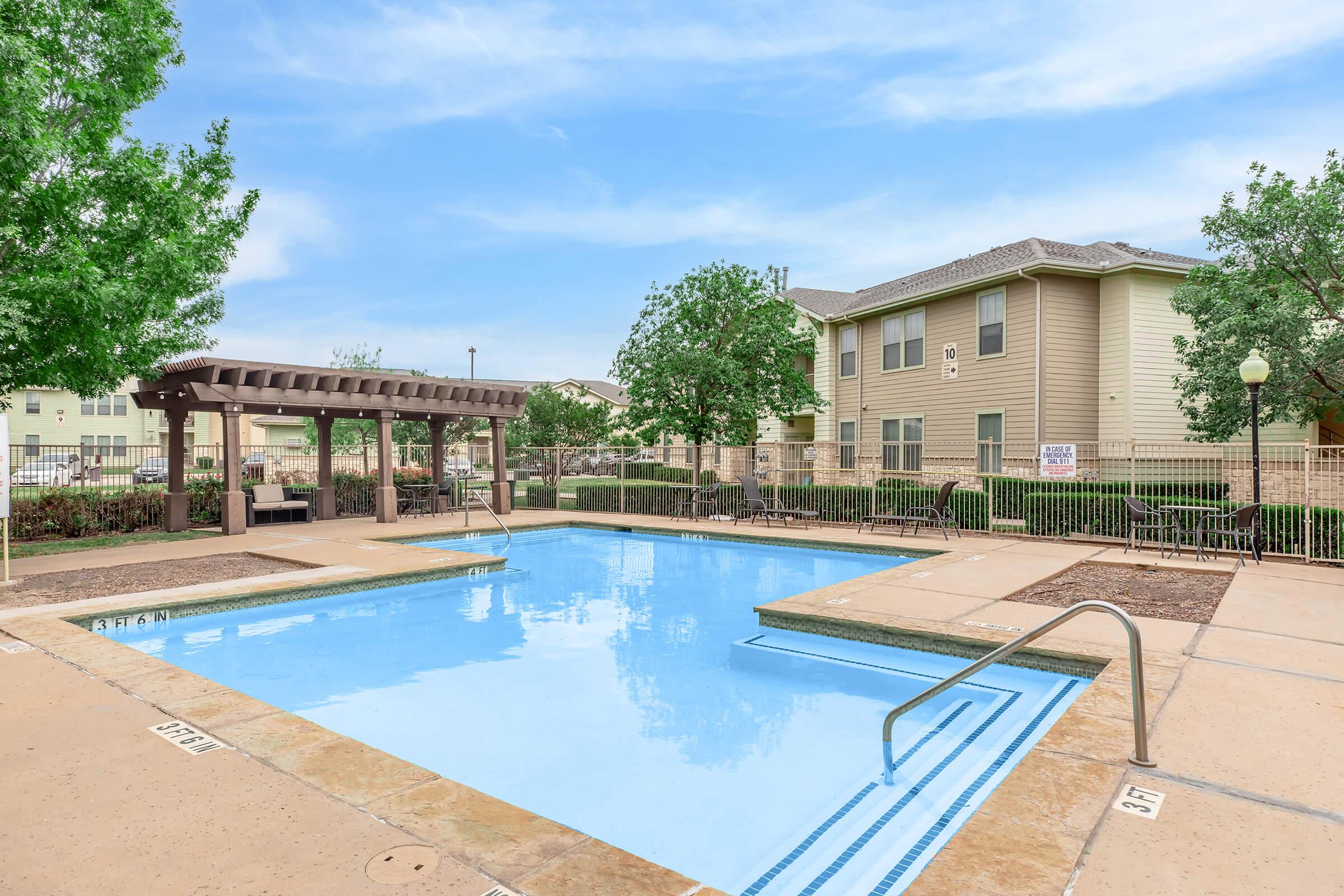 A clean, blue swimming pool surrounded by a concrete patio, with a shaded pergola area nearby. Lush green trees and landscaped shrubs add to the ambiance, with a multi-story residential building visible in the background under a partly cloudy sky.