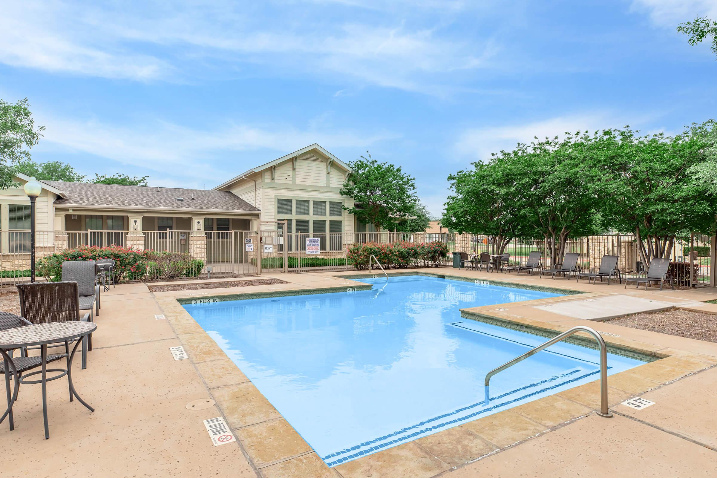 A serene swimming pool surrounded by lounge chairs and greenery. The pool area features a light blue pool with a railing, a fenced building in the background, and well-maintained landscaping, creating a relaxing outdoor atmosphere.