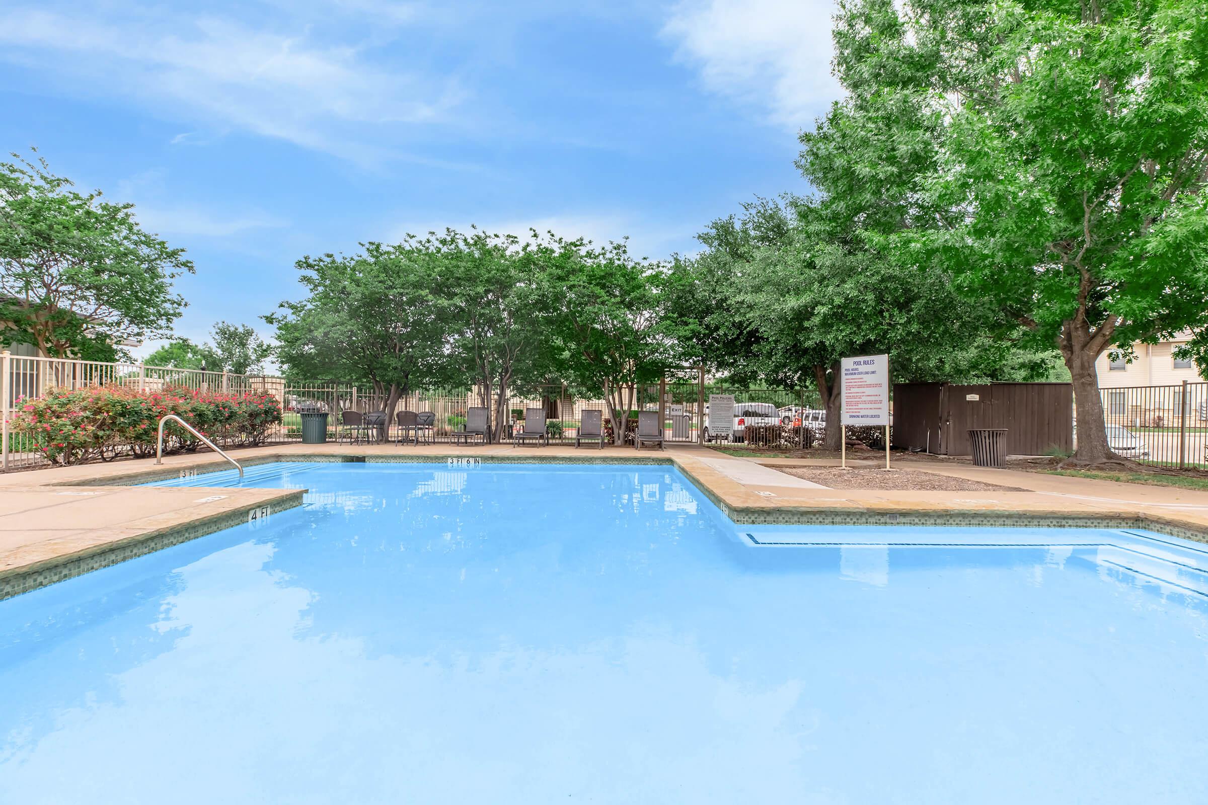 A clear blue swimming pool surrounded by green trees and shrubs, with lounge chairs placed nearby. The area is well-maintained and features a sign near the pool. The sky is bright and partly cloudy, creating a serene outdoor atmosphere.