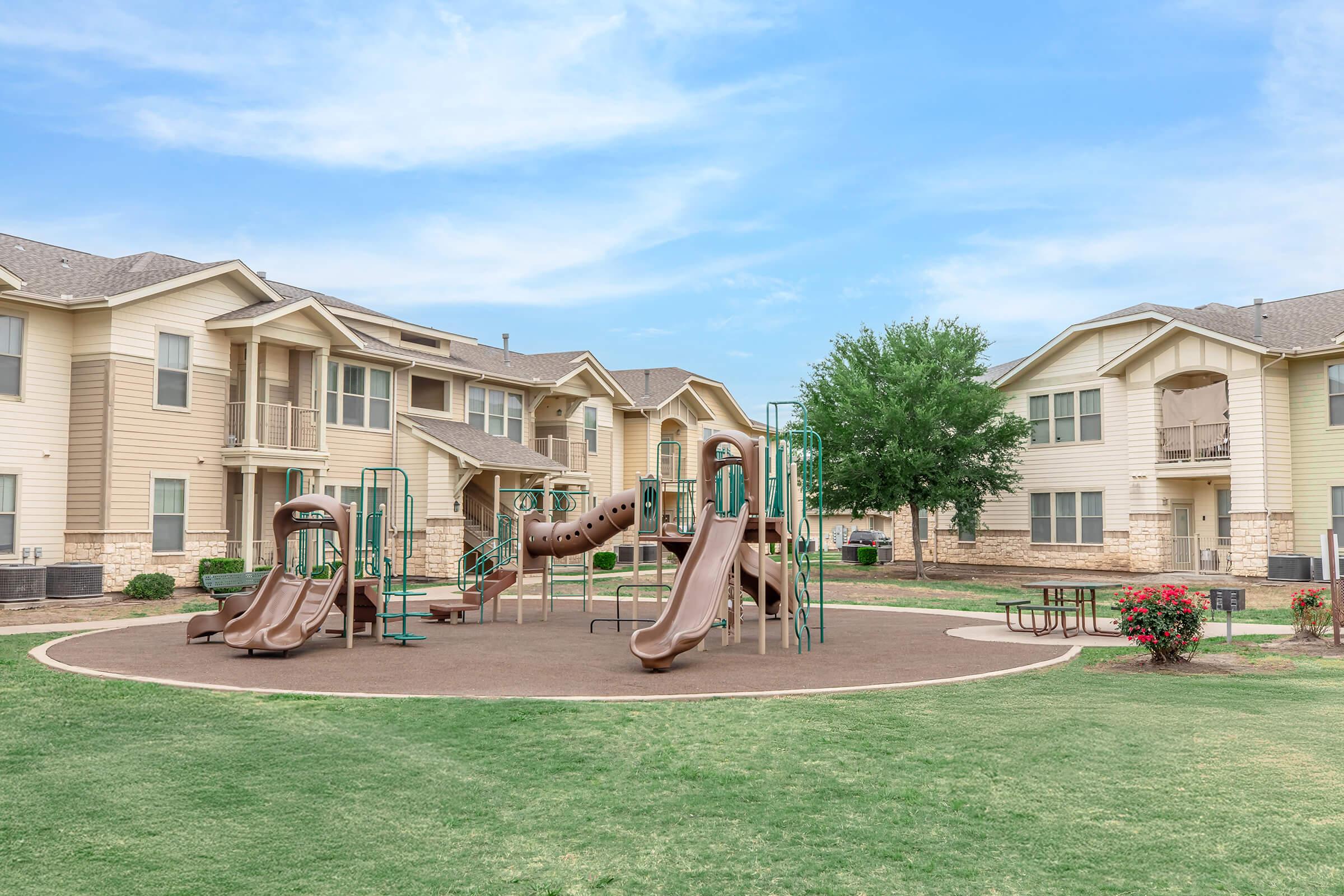 A playground with various slides and climbing structures surrounded by green grass and flower bushes, located in an apartment complex. Several buildings with balconies are visible in the background under a clear blue sky.