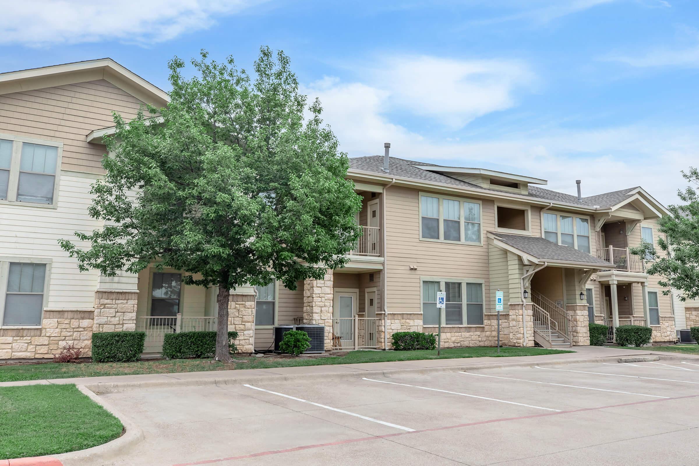 A multi-unit residential building featuring modern architecture with light-colored siding. The structure has multiple balconies, green landscaping, and a parking area in front. Trees provide shade, and the sky is partly cloudy, creating a pleasant atmosphere.
