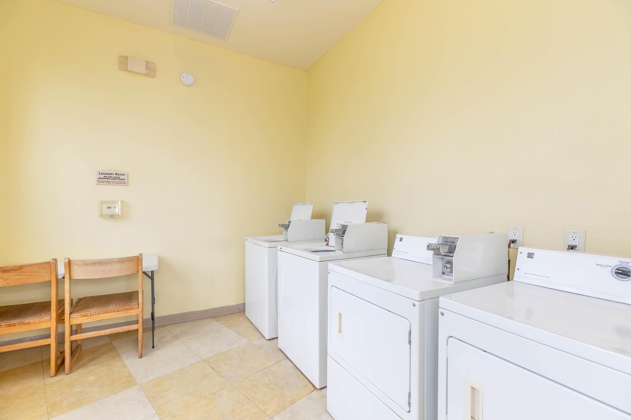 A laundry room featuring two rows of white washing machines and dryers against a light yellow wall. There are two wooden chairs positioned nearby for seating. The floor is tiled with light-colored tiles, creating a clean and functional space for laundry tasks.