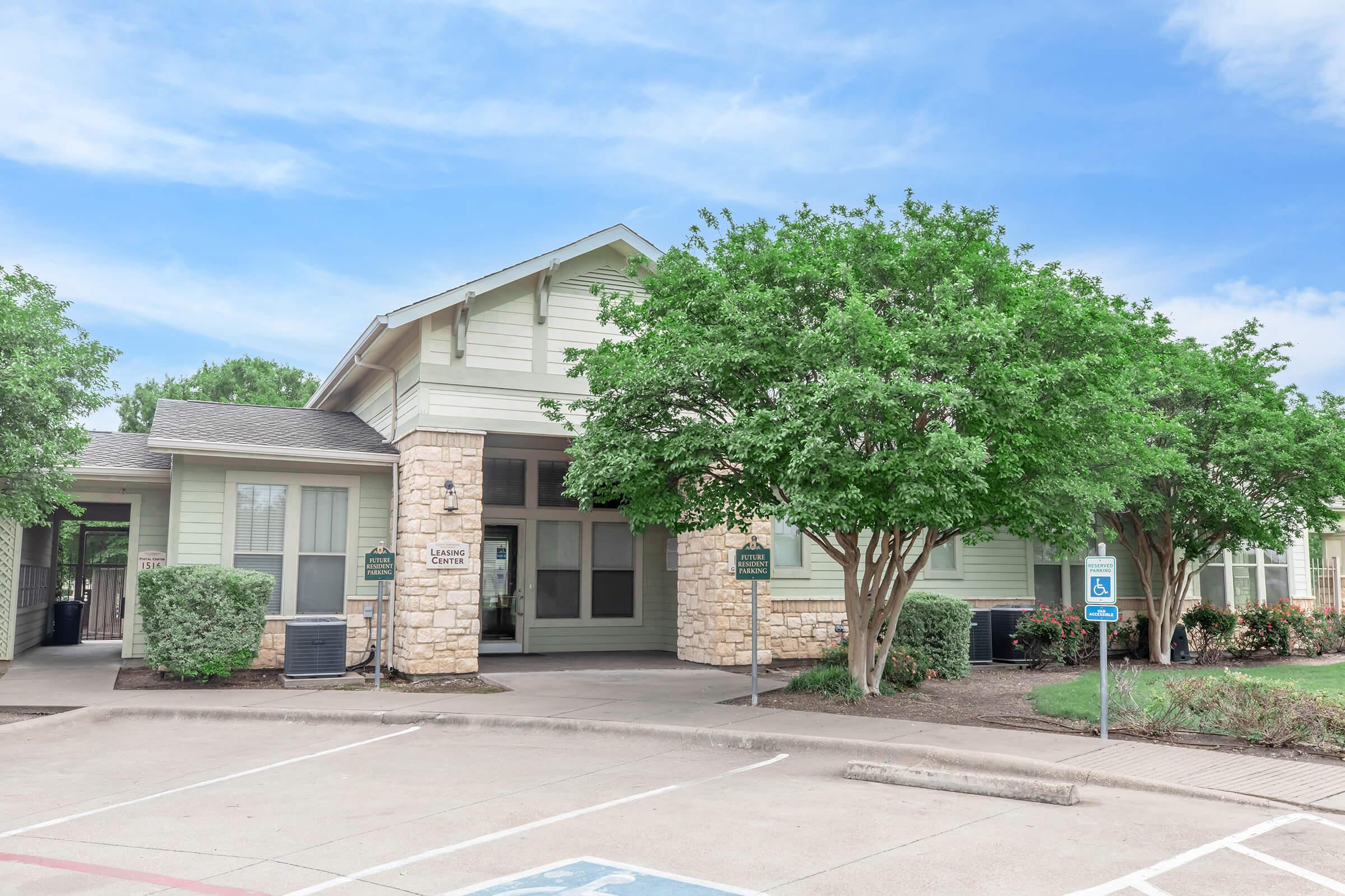 A light green building with stone accents houses the leasing center. It features large windows and is surrounded by well-maintained trees and landscaping. The parking lot has designated spots, including a handicapped space, and the sky is clear with a few clouds.