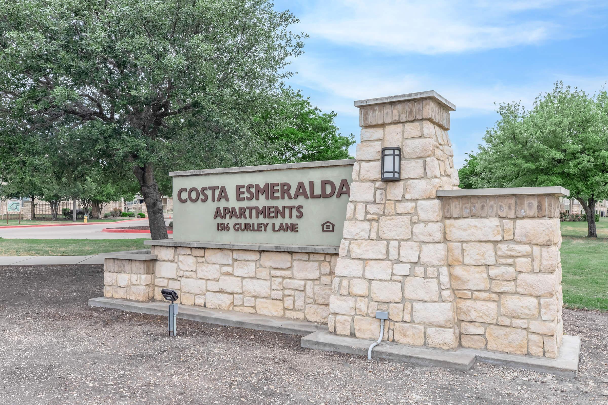 Stone sign for Costa Esmeralda Apartments located at 1516 Gurley Lane. The sign features a modern design with landscaping and trees in the background, set in a residential area.