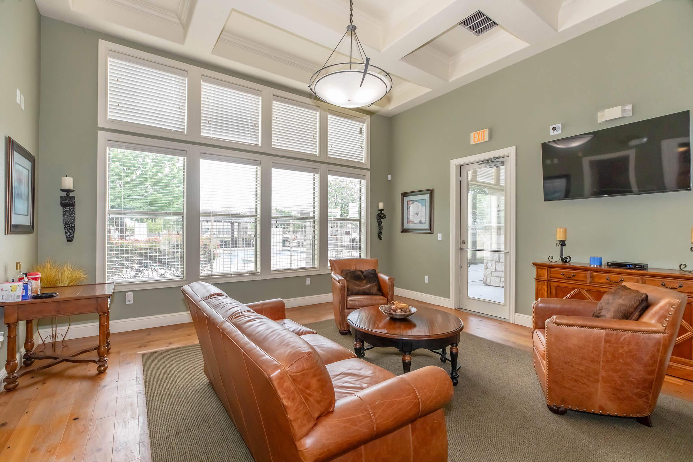 A cozy living room with large windows allowing natural light, featuring a circular wooden coffee table and brown leather sofas. A wooden sideboard holds decorative items, while a flat-screen TV is mounted on the wall. The room has a stylish chandelier and neutral-colored walls, creating a warm atmosphere.