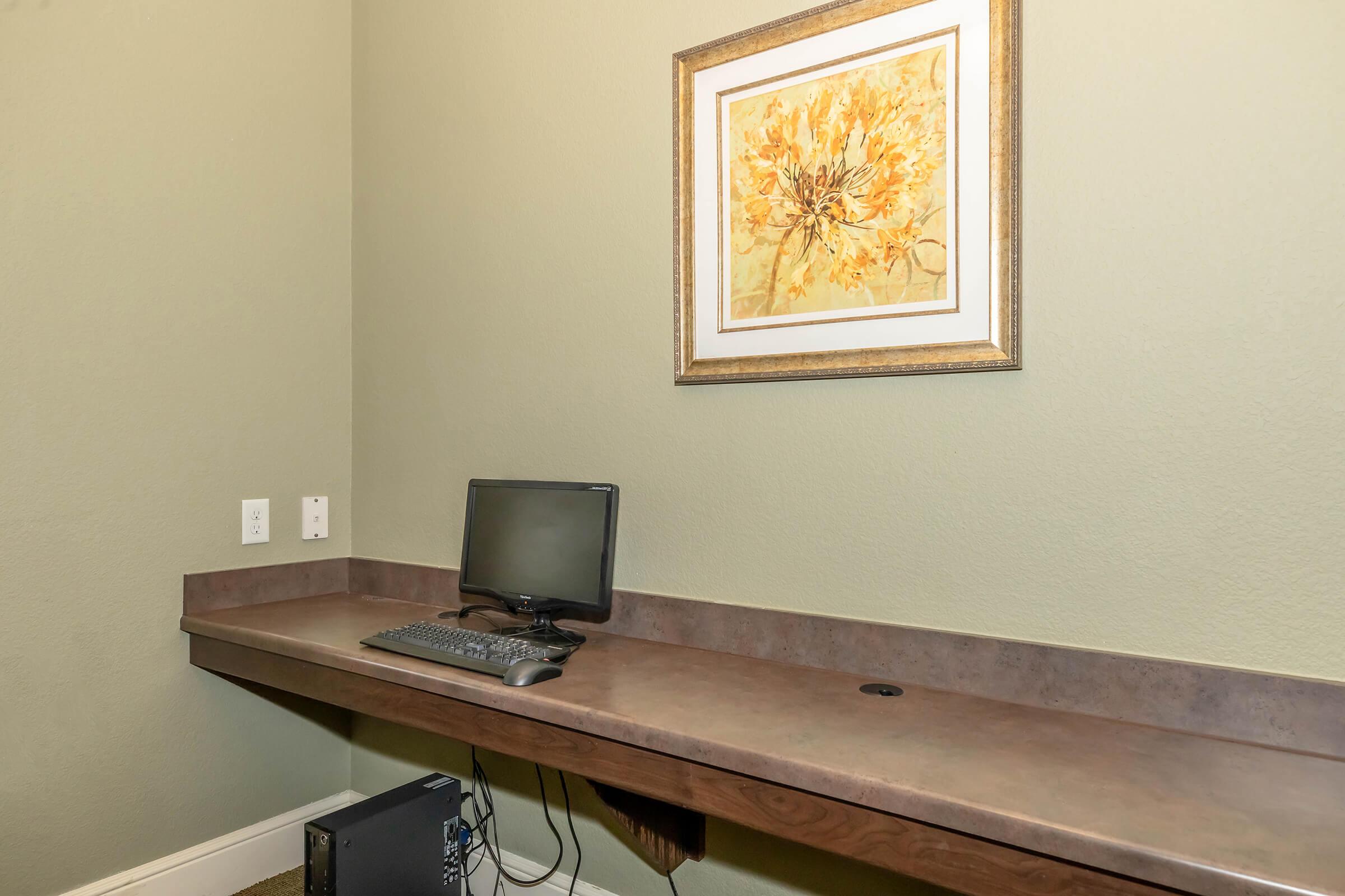 A small office space featuring a computer monitor with a keyboard, situated on a brown desk against a green wall. Above the desk, there is a framed artwork depicting yellow flowers, adding a touch of color to the environment. The overall setup is simple and functional.