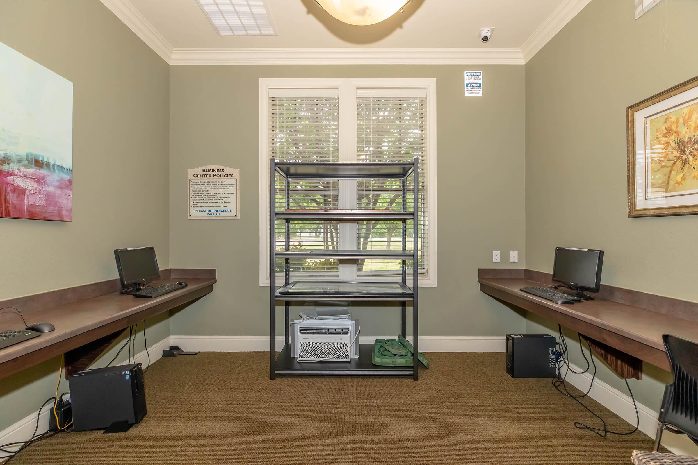 A small office space with two wooden desks equipped with computers on either side. In the center, there is a metal shelving unit. The walls are painted green, and there are windows with blinds letting in natural light. A sign is visible on the wall, and decorative artwork hangs nearby.