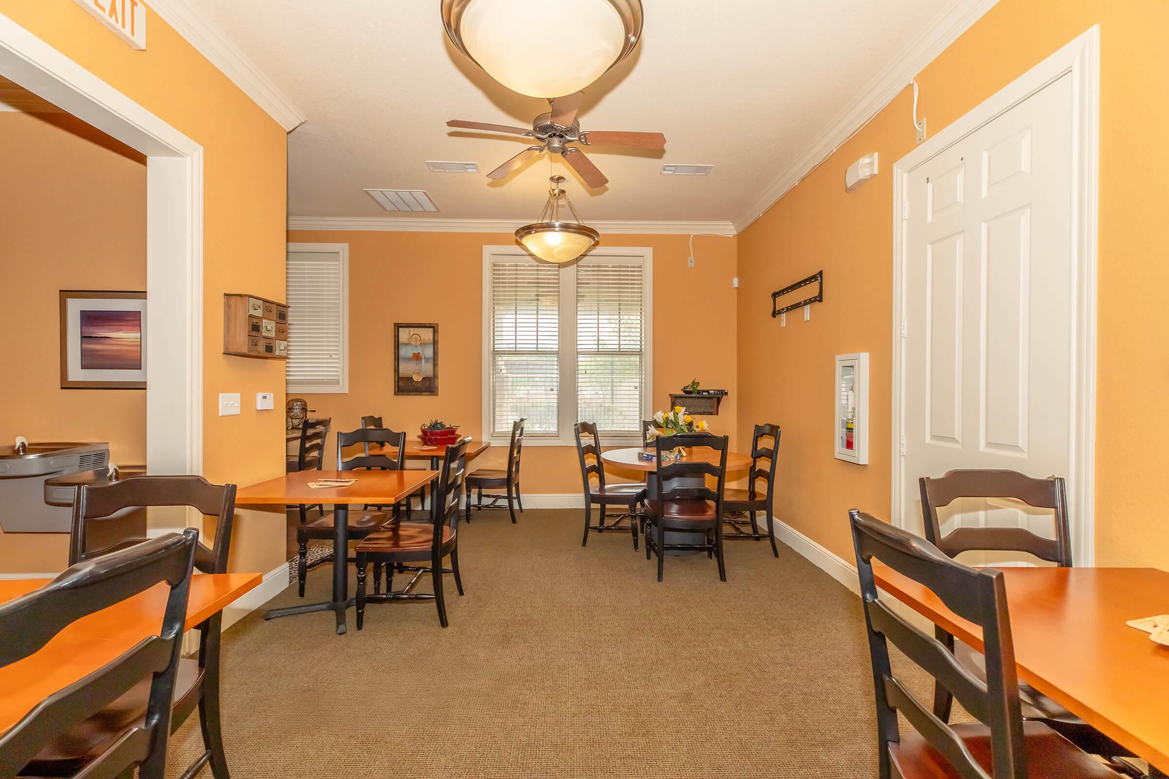 Interior of a cozy dining area featuring orange walls and wooden tables with black chairs. Ceiling fans and warm lighting create a welcoming atmosphere. Windows let in natural light, and a decorative touch is added with potted plants. The space is tidy and well-organized, suitable for gatherings or casual dining.