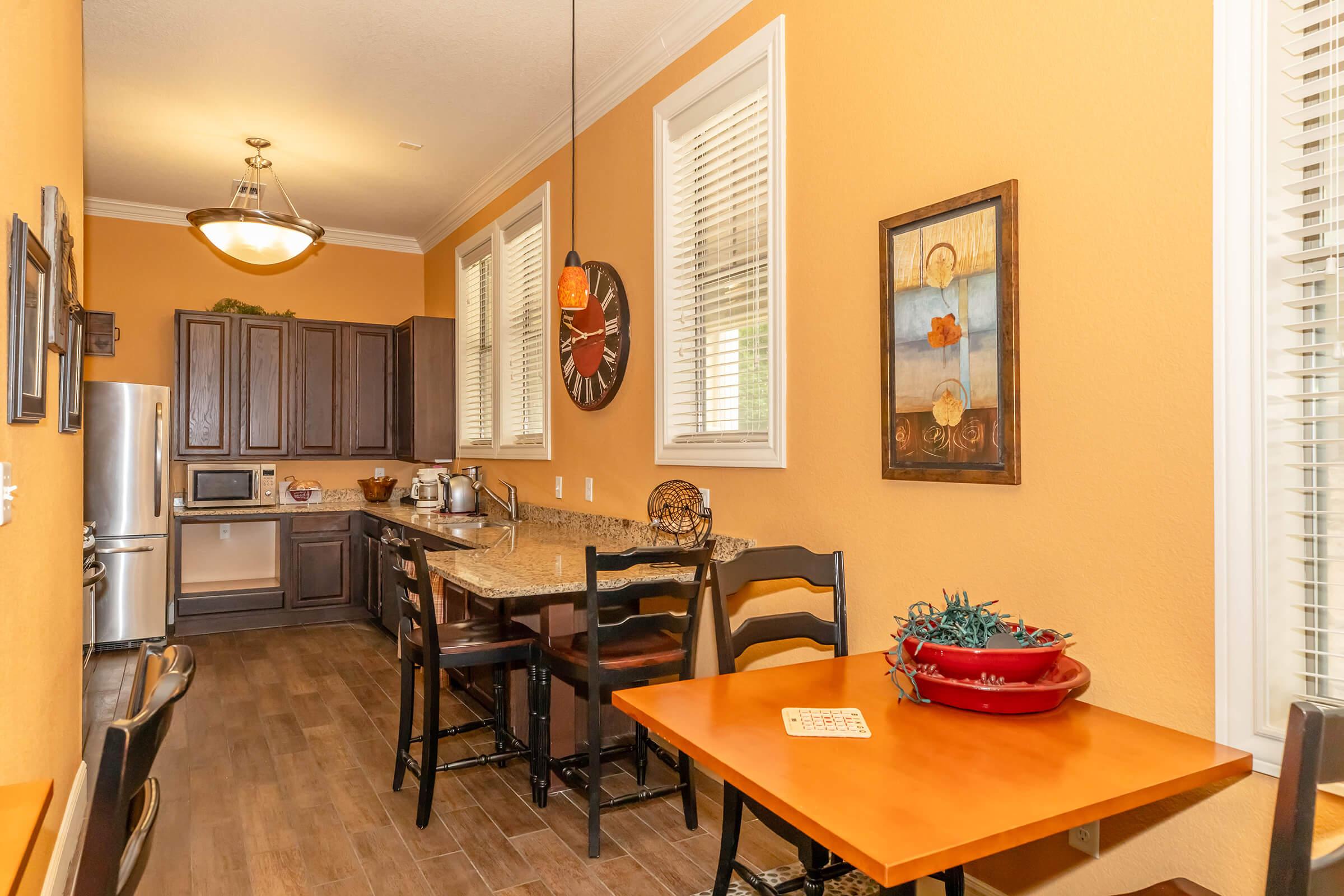 Modern kitchen with warm orange walls, featuring wooden cabinets, stainless steel appliances, and a granite countertop. A dining area with a small orange table and chairs is visible. Decorative wall art and natural light from windows enhance the inviting atmosphere.