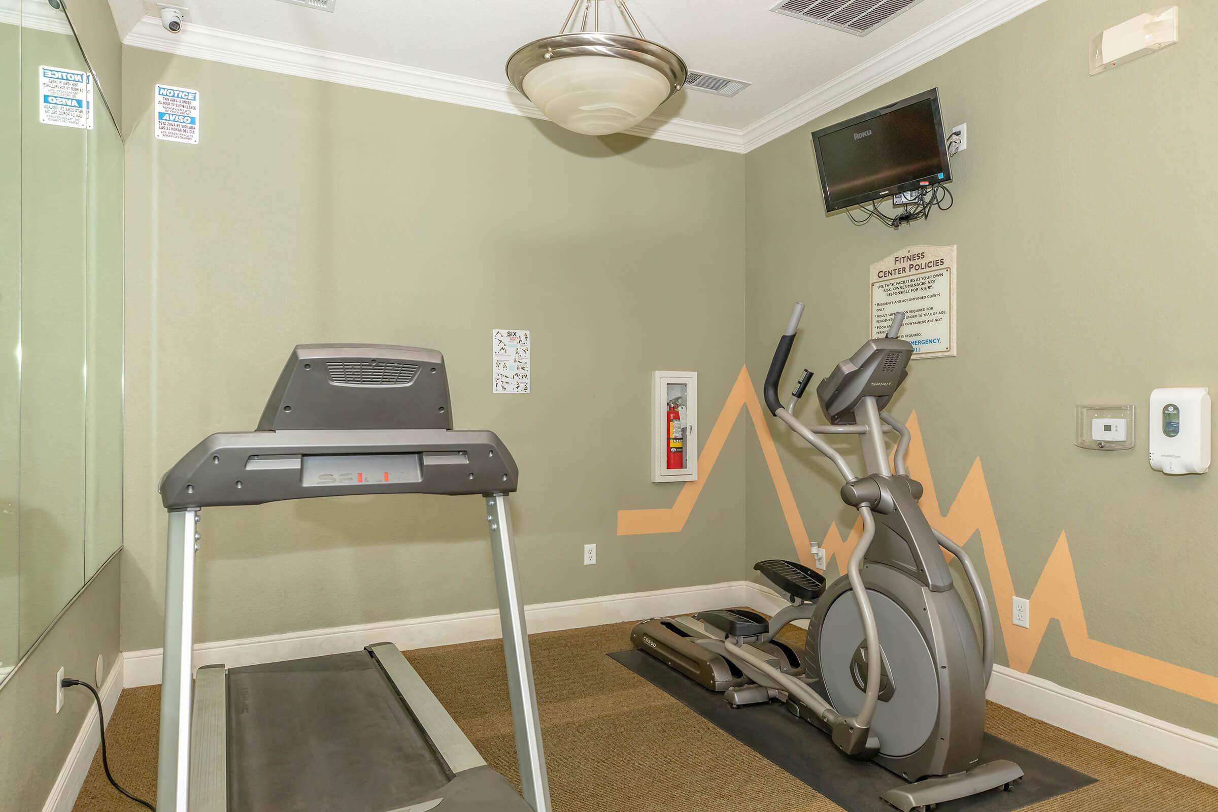 A small fitness room featuring a treadmill and an elliptical machine. The walls are painted in a muted green color with minimal decor. There's a wall-mounted TV, a fire extinguisher on the wall, and gym instructions visible on a nearby sign.
