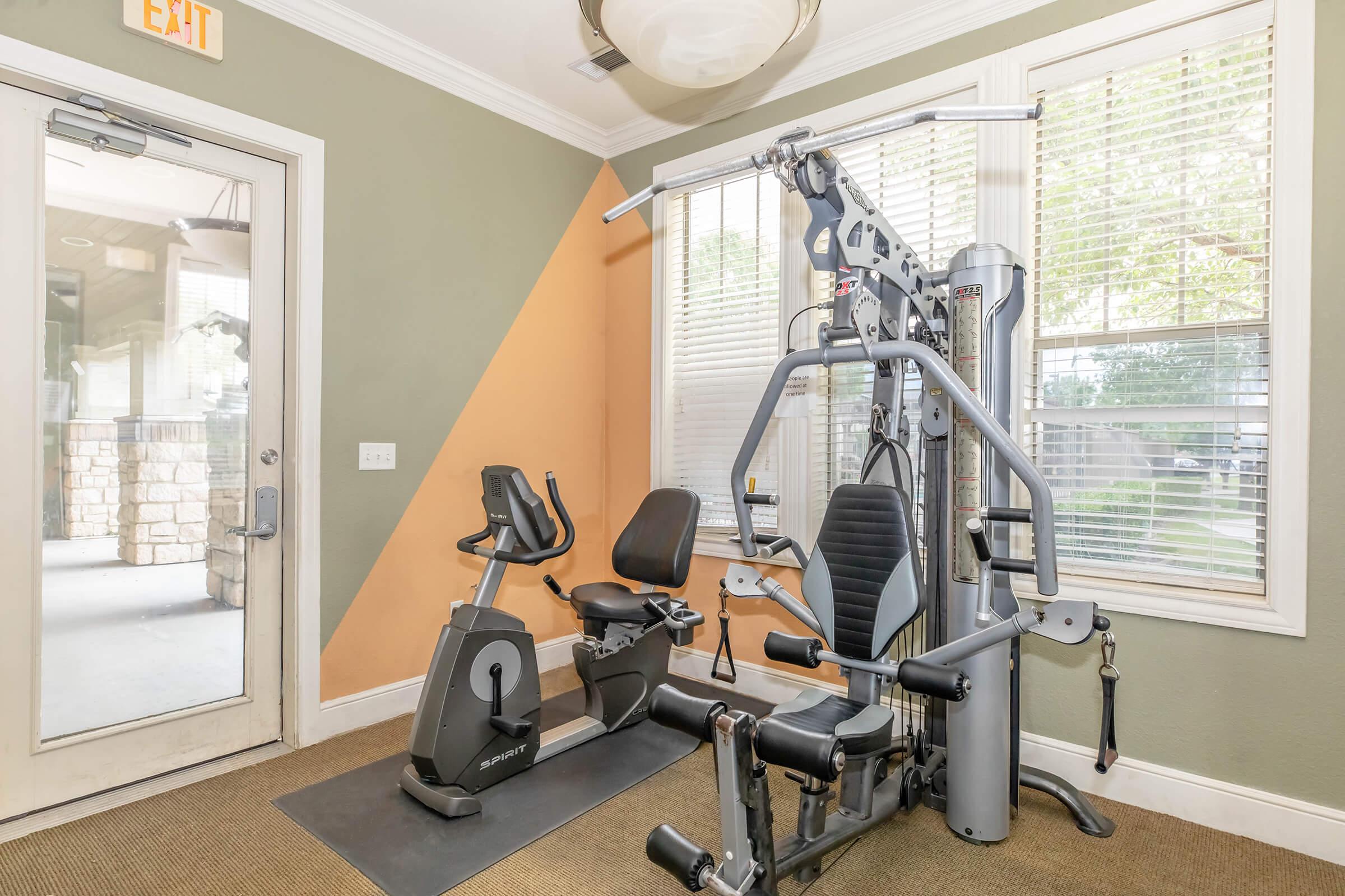 A brightly lit fitness room featuring a multi-gym machine, a recumbent exercise bike, and large windows allowing natural light. The walls are painted in green and orange, creating a vibrant atmosphere for workouts. A door leads to an outdoor area.