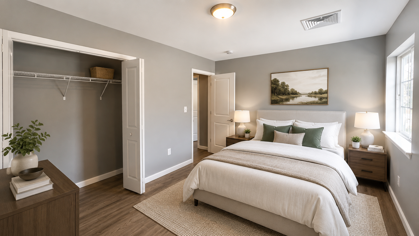 A modern bedroom featuring a neatly made bed with white linens and green throw pillows. There are two bedside tables with lamps and a decorative plant. A large window allows natural light, and an art piece hangs on the wall. An empty closet space is visible on the left, adding to the room's minimalistic aesthetic.
