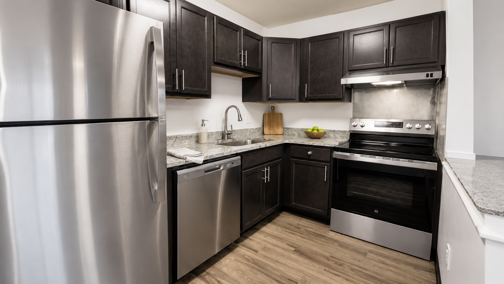 Modern kitchen featuring dark wood cabinets, stainless steel appliances including a refrigerator, dishwasher, and oven. The countertop is grey with a small sink, and a wooden cutting board sits against the wall. The floor is light wood, creating a warm, inviting atmosphere.