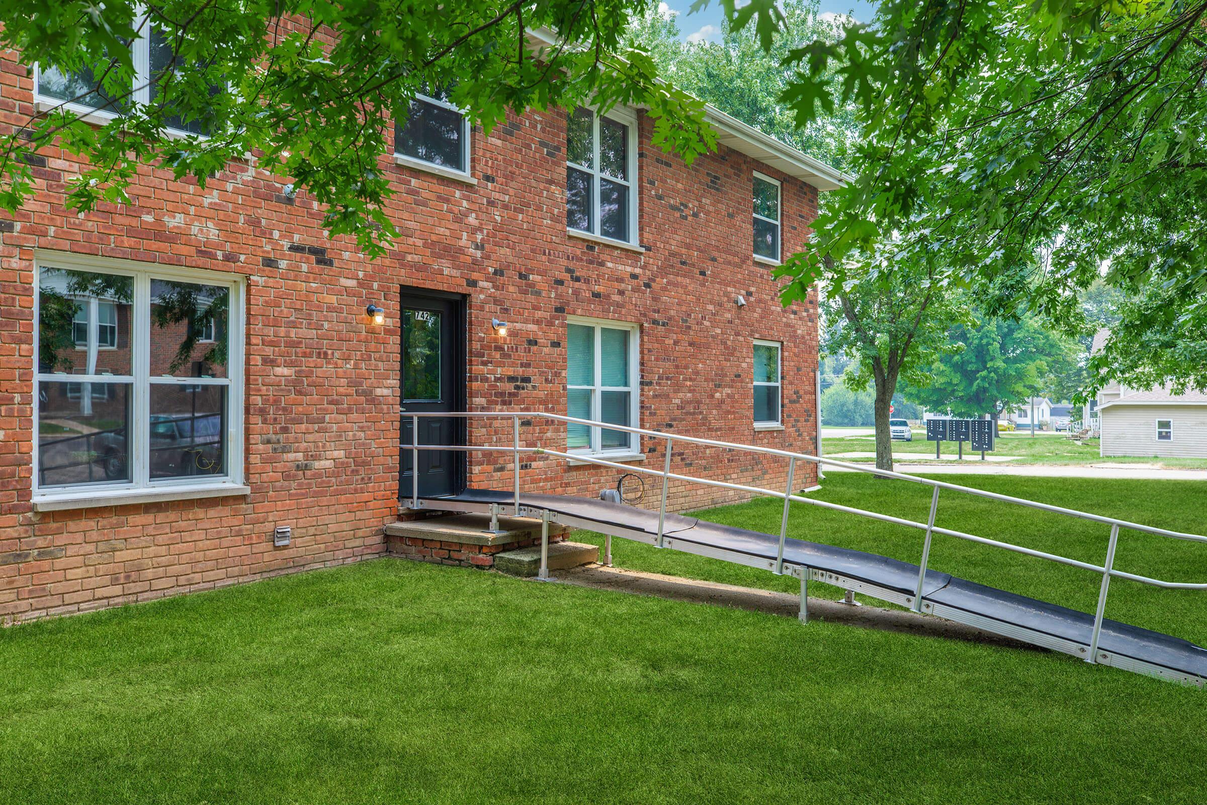 A brick building with large windows and a wheelchair ramp leading to the entrance. The landscape features well-maintained green grass and trees offering shade, with a clear view of nearby buildings in the background. The setting appears welcoming and accessible.