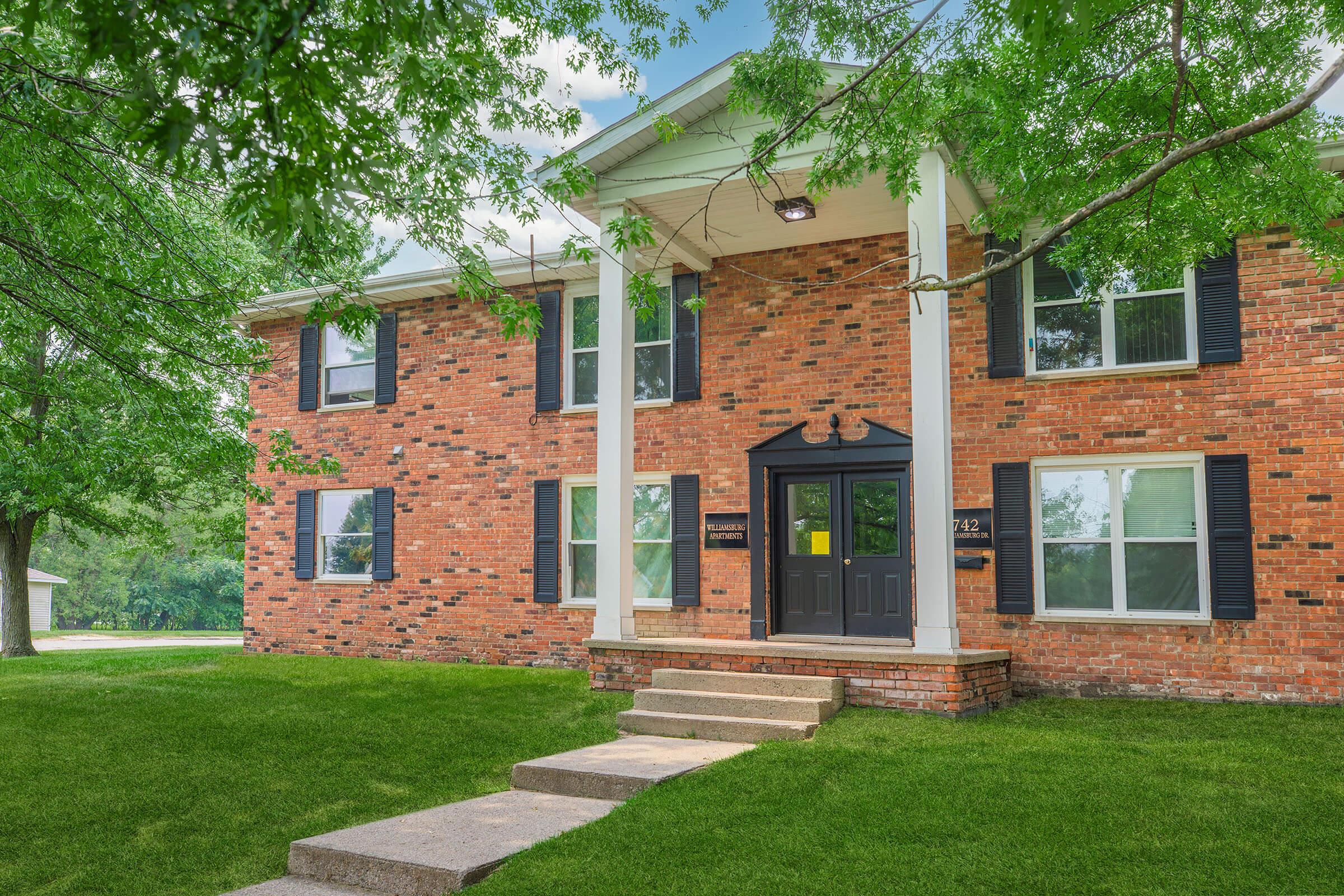 A brick two-story house surrounded by green trees and grass. The house features large windows with black shutters, a front porch with steps leading up to a black door, and decorative elements above the door. The sky is clear and sunny, adding to the bright, inviting atmosphere.