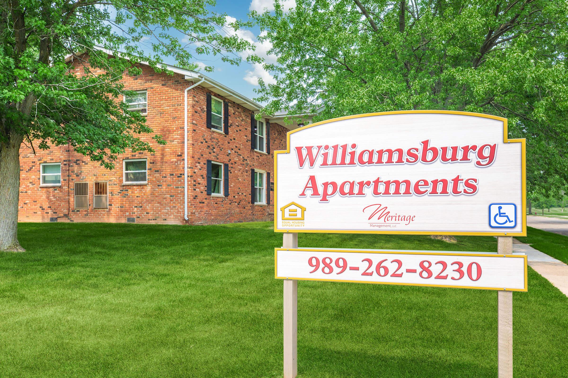 Sign for Williamsburg Apartments featuring the phone number 989-262-8230. The brick building behind the sign is surrounded by greenery and has multiple windows. The ground is well-maintained, with grass and trees providing a pleasant atmosphere.