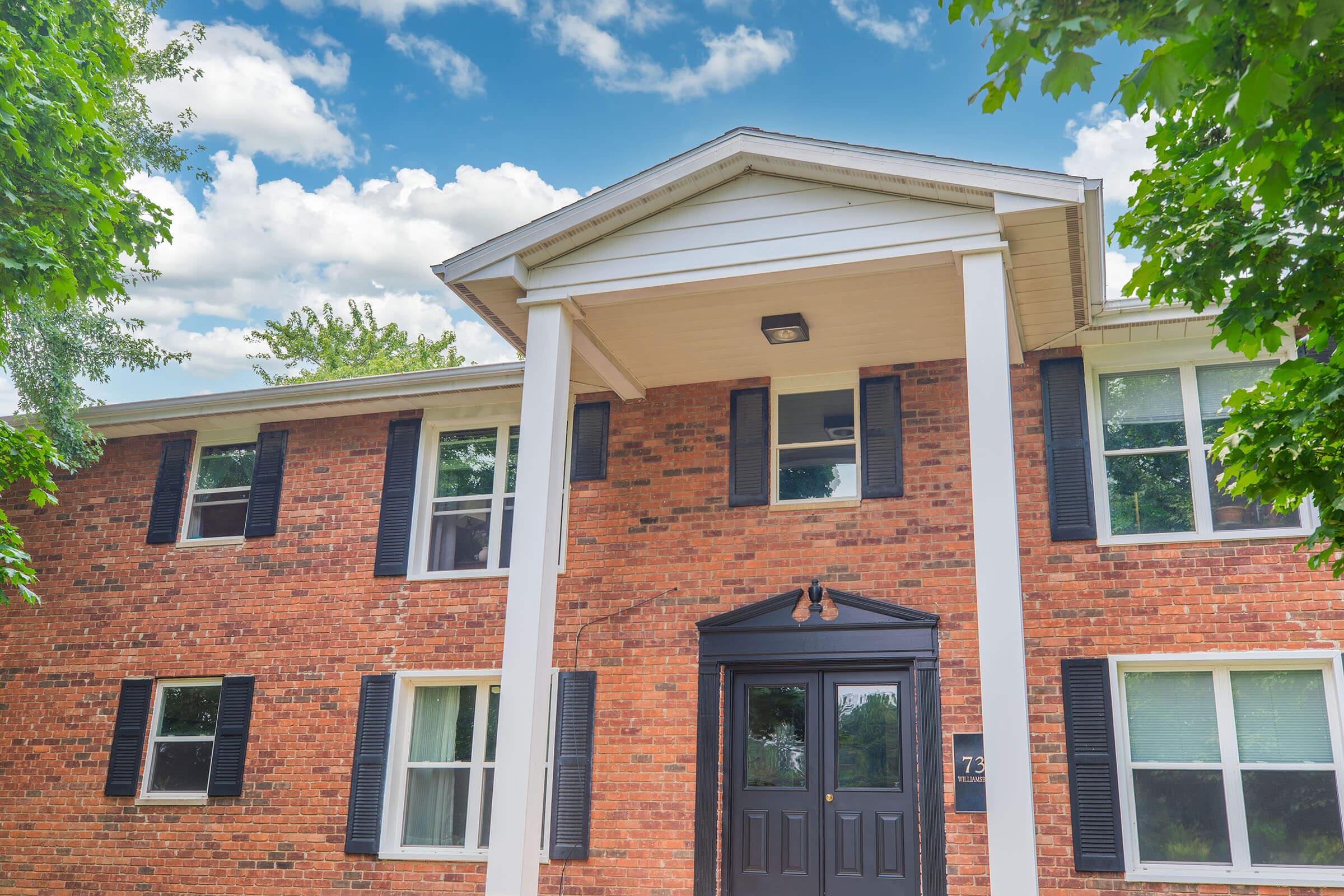 A two-story brick building with black shutters and a central entrance featuring a dark door. The façade is adorned with white columns and the sky is clear with scattered clouds, surrounded by green trees.