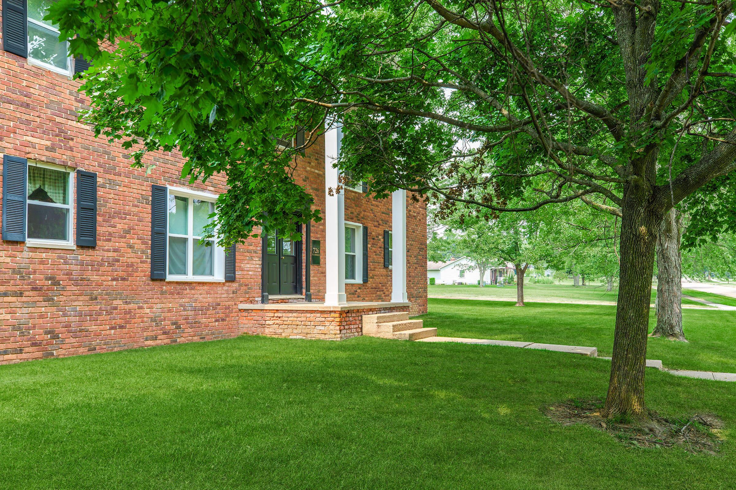 A well-maintained brick building with white trim and black shutters is shown. The entrance features a small set of steps leading to a door, surrounded by a lush green lawn and trees. The scene is bright and inviting, showcasing a peaceful and residential neighborhood atmosphere.