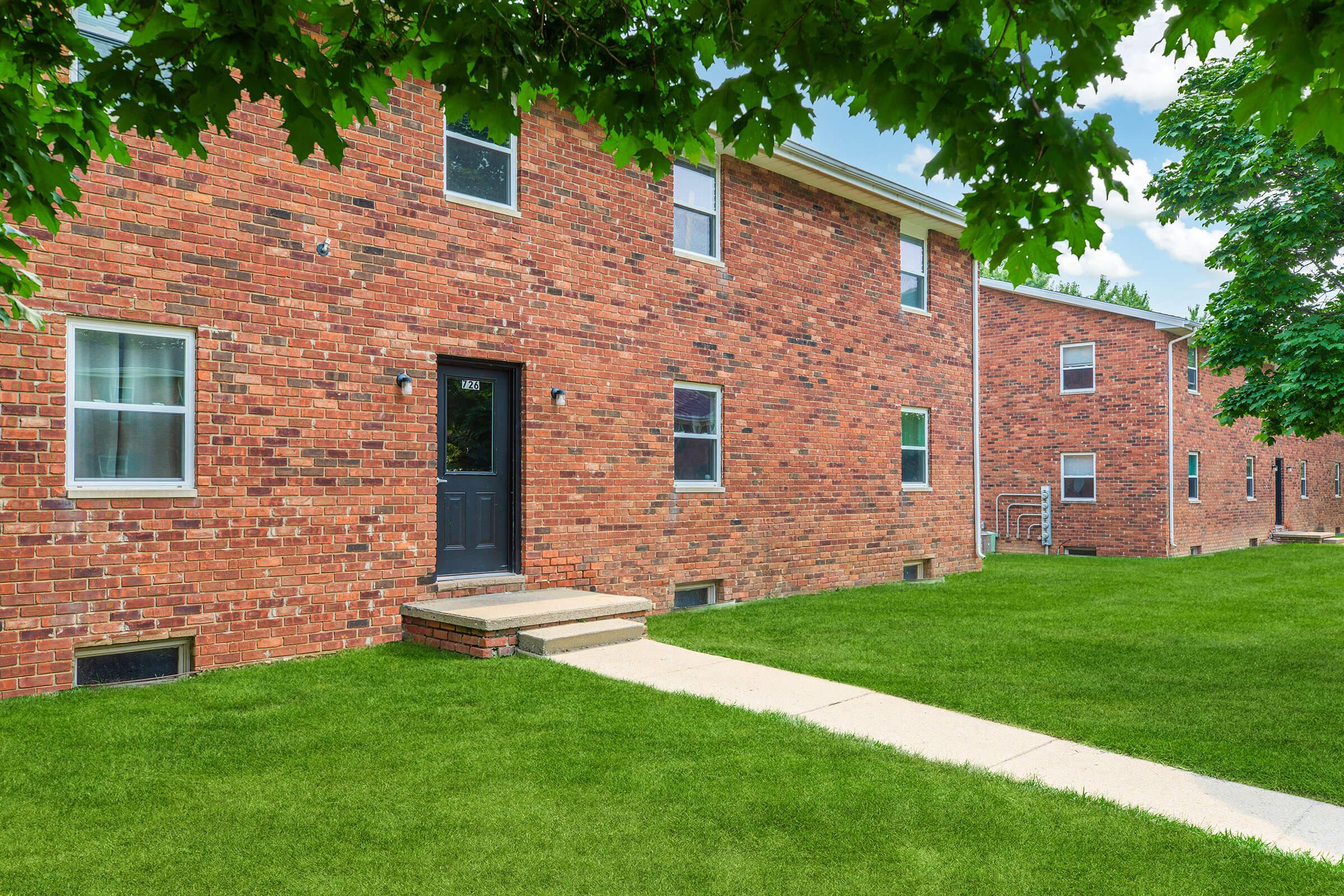 A brick apartment building with a dark front door and five windows, surrounded by green grass. A concrete walkway leads to the entrance, and trees provide shade around the property. The sky is partly cloudy, adding to the serene atmosphere of the residential area.