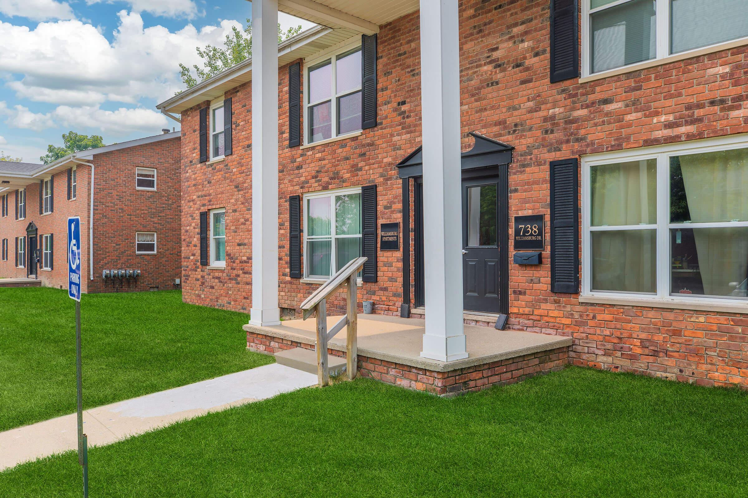 A brick apartment building with white trim and a black front door, featuring a sloped walkway leading up to the entrance. The lawn is well-maintained with green grass, and there is a blue handicap accessibility sign nearby. The sky is partly cloudy, adding to the pleasant ambiance of the setting.
