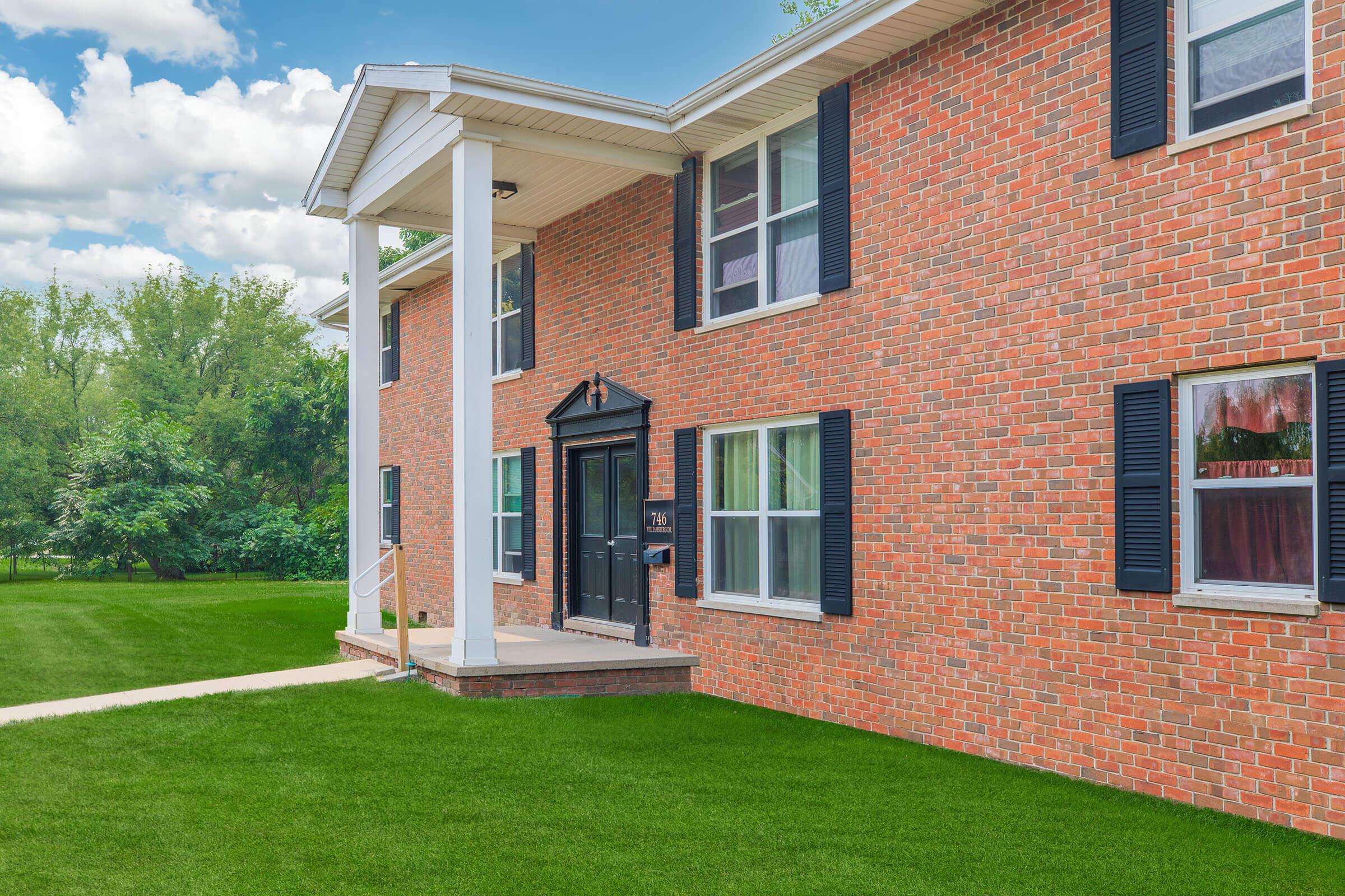 A brick building with large windows, black shutters, and a central entrance featuring a dark door. The facade is complemented by white columns. The surrounding area has well-maintained green grass and trees in the background under a partly cloudy sky.