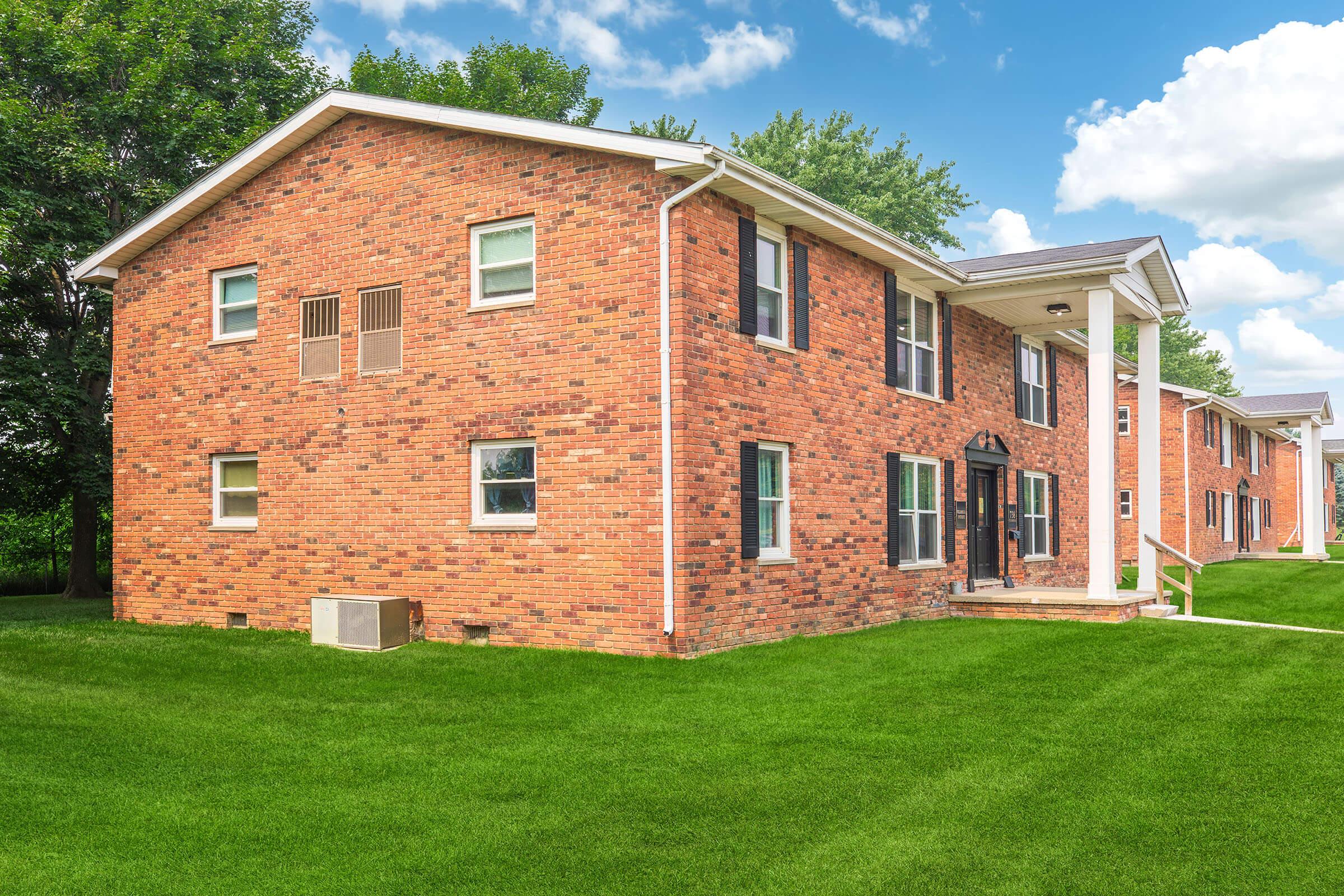 A two-story brick apartment building with large windows and a front entrance. The building is surrounded by a well-maintained green lawn, and there are trees and blue skies in the background, creating a sunny and inviting atmosphere.