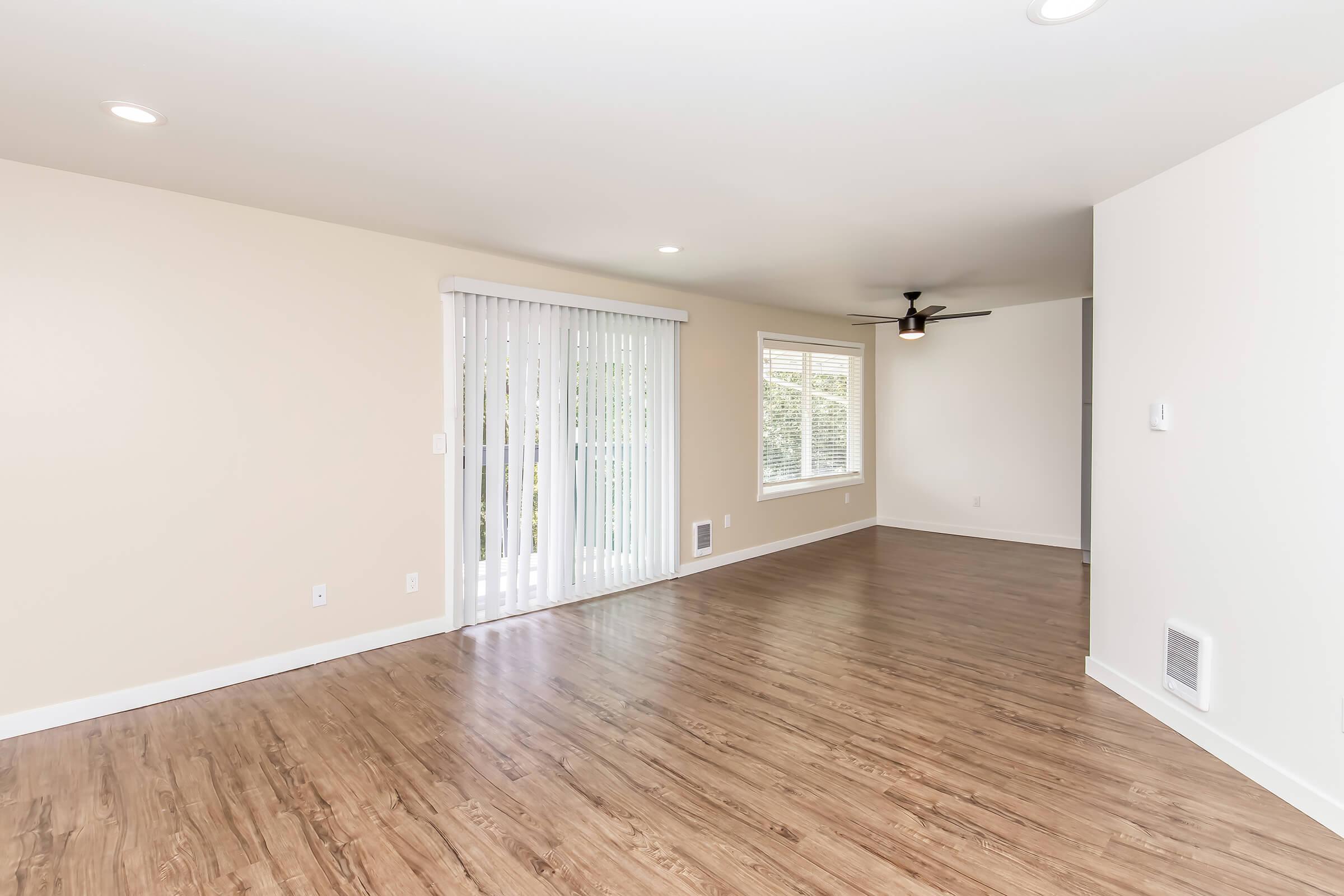 A spacious living room with light wood laminate flooring, beige walls, and large windows. The room features sliding glass doors leading to a patio, a ceiling fan, and natural light illuminating the interior.