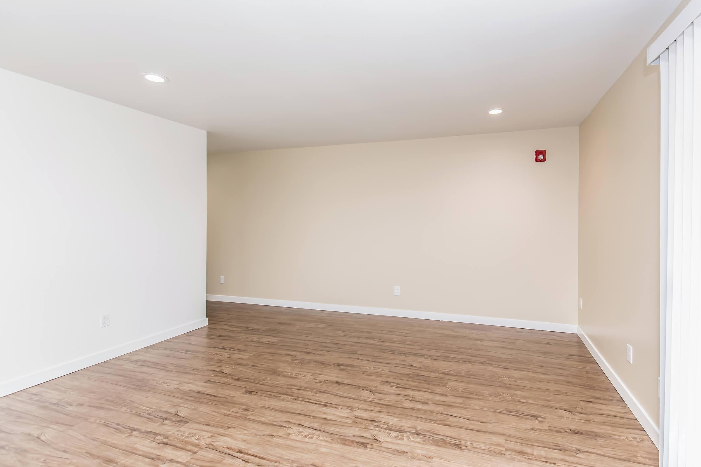 A spacious, empty room with light-colored walls and a polished wooden floor. Two ceiling lights provide illumination. There are no furnishings, creating a minimalist feel. A small red wall alarm is visible on one wall, and a sliding door is positioned on the left side.