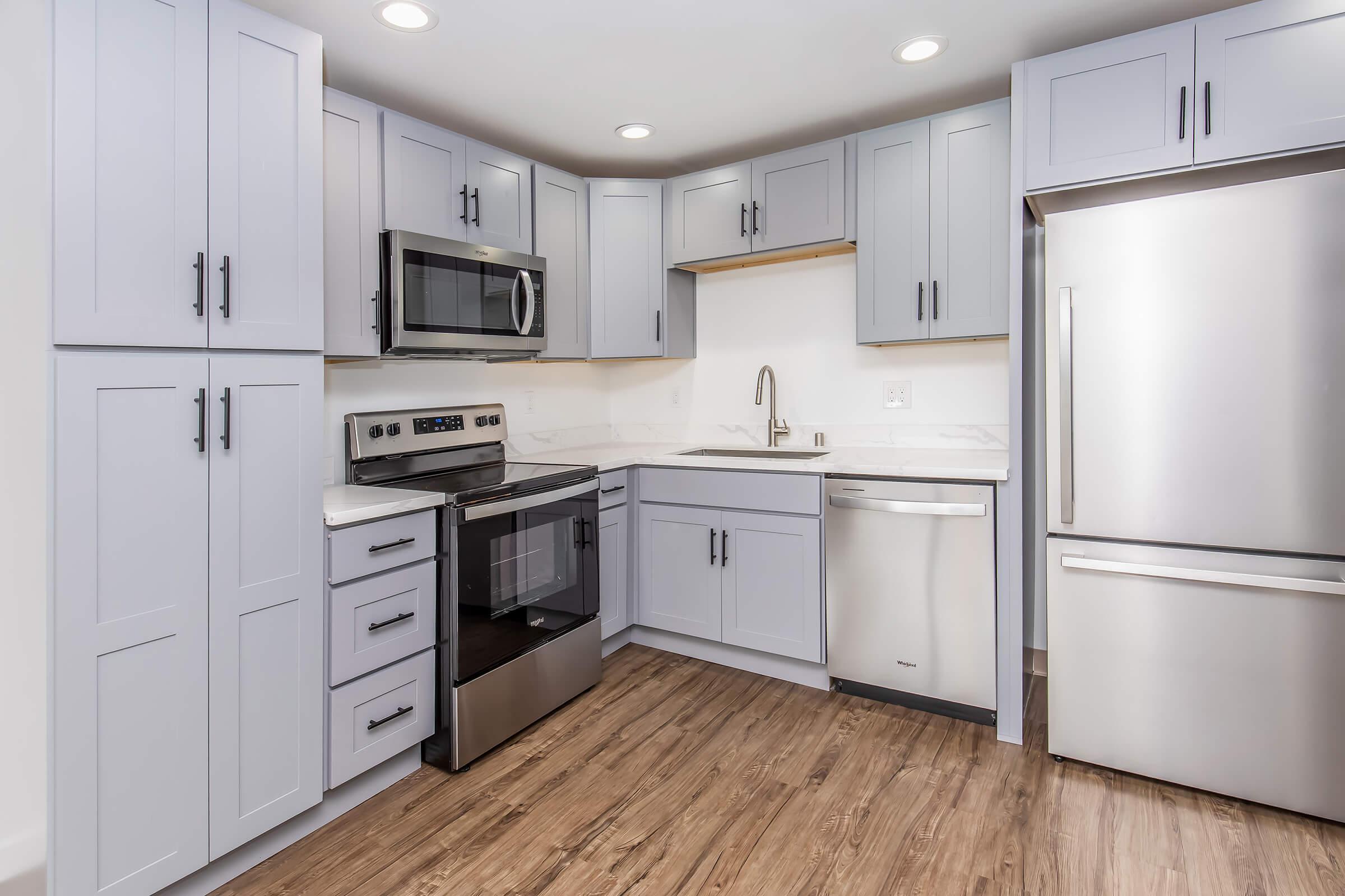 Modern kitchen featuring light gray cabinets, a stainless steel refrigerator, an oven, and a microwave. The countertops are white with subtle veining, and there is a silver sink with a modern faucet. The flooring is a wood-like laminate that adds warmth to the space. The kitchen is well-lit with recessed lighting.